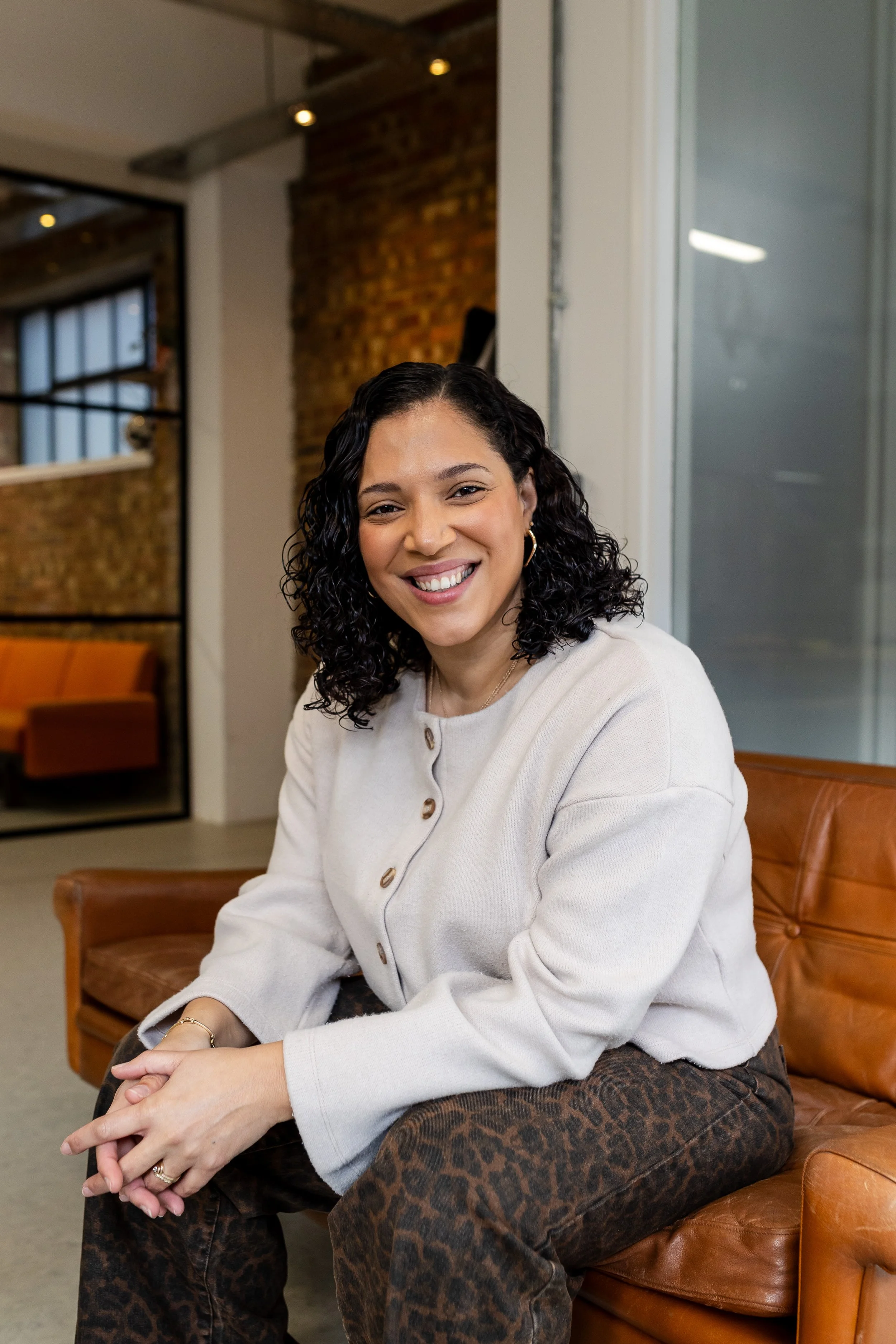 A woman with curly black hair smiling, sitting on a leather chair in an indoor setting with exposed brick wall and large window.