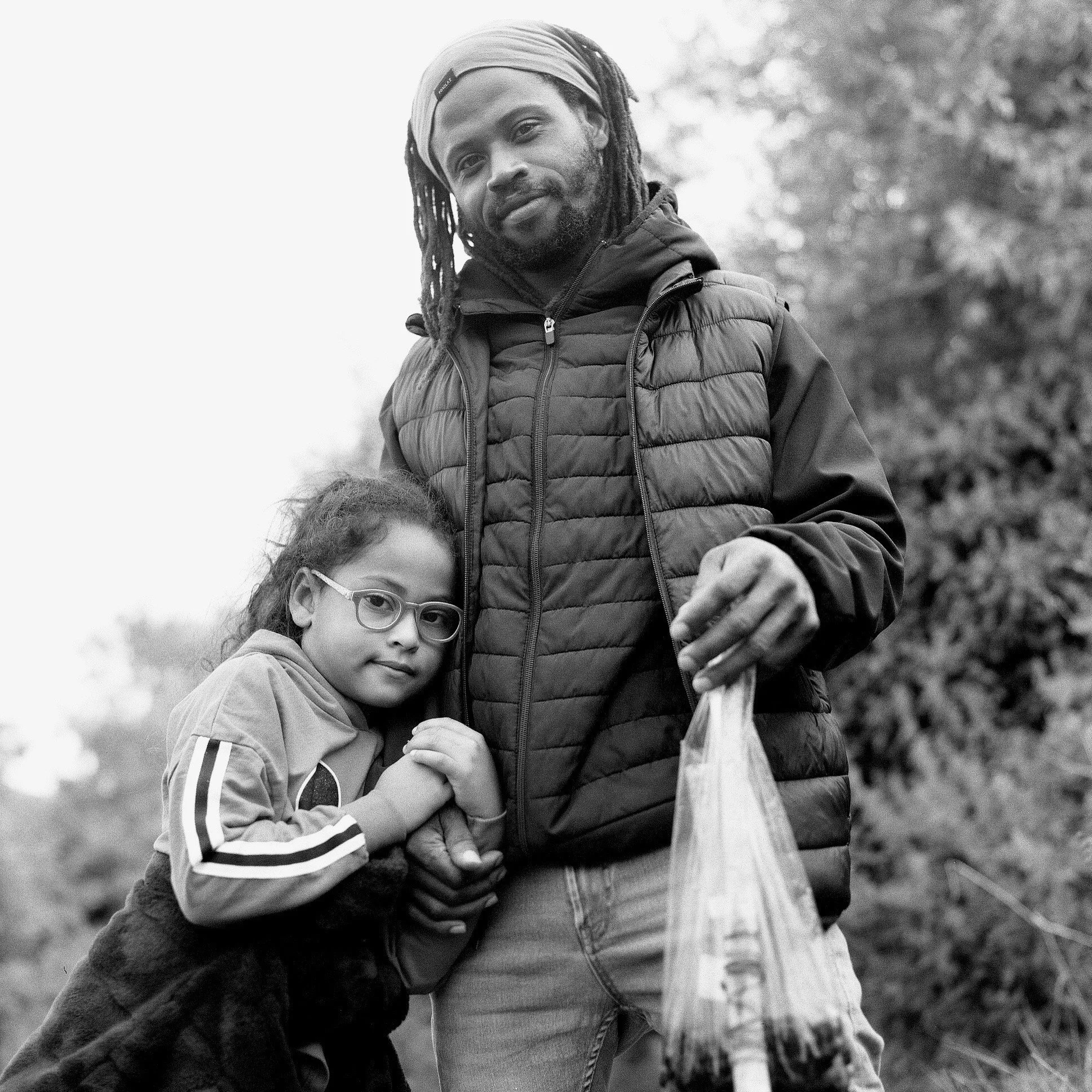 A black-and-white photo of a man with long dreadlocks wearing a head wrap and a puffer vest, standing outdoors near the trees. A girl with glasses and curly hair, wearing a striped jacket, is in front of him, holding his hand.