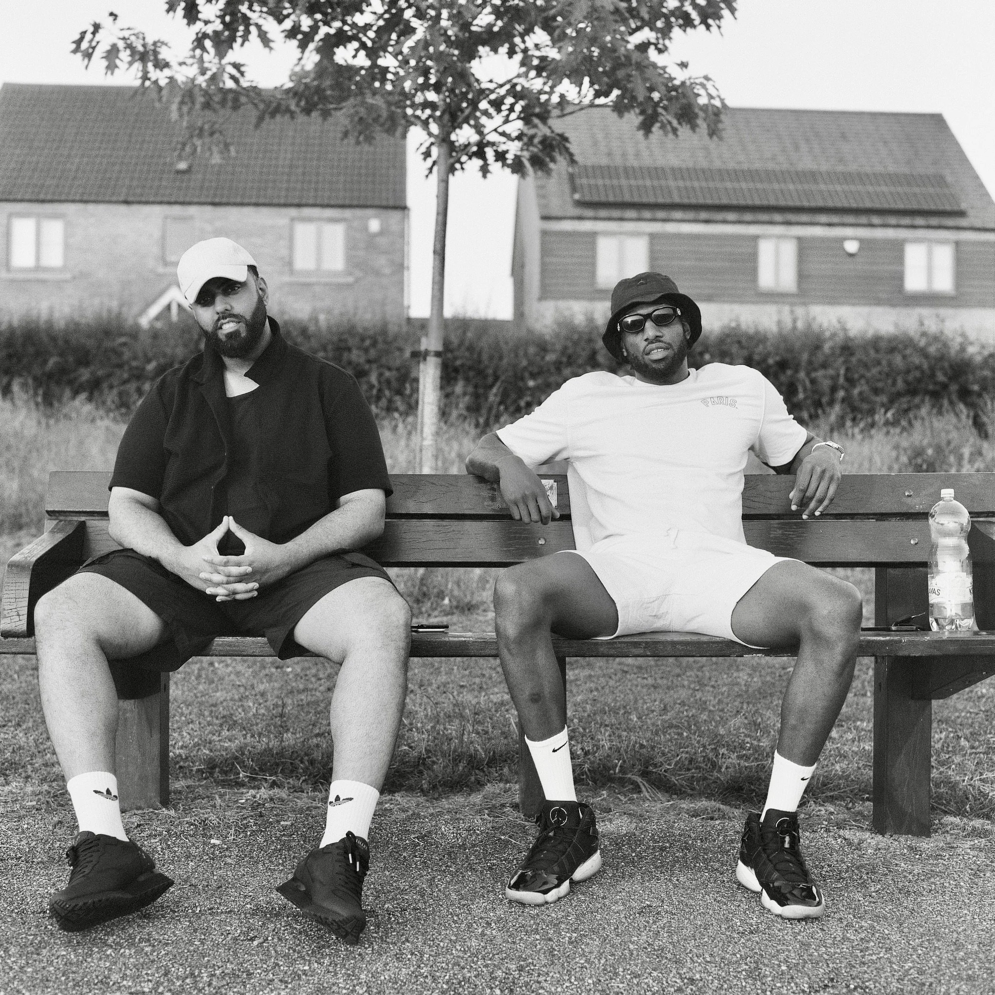 Two men sitting on a wooden bench outdoors, with trees and residential buildings in the background. One man is wearing a cap and dark clothing, the other wearing sunglasses, a bucket hat, and a white t-shirt. They both have athletic shoes and socks, 