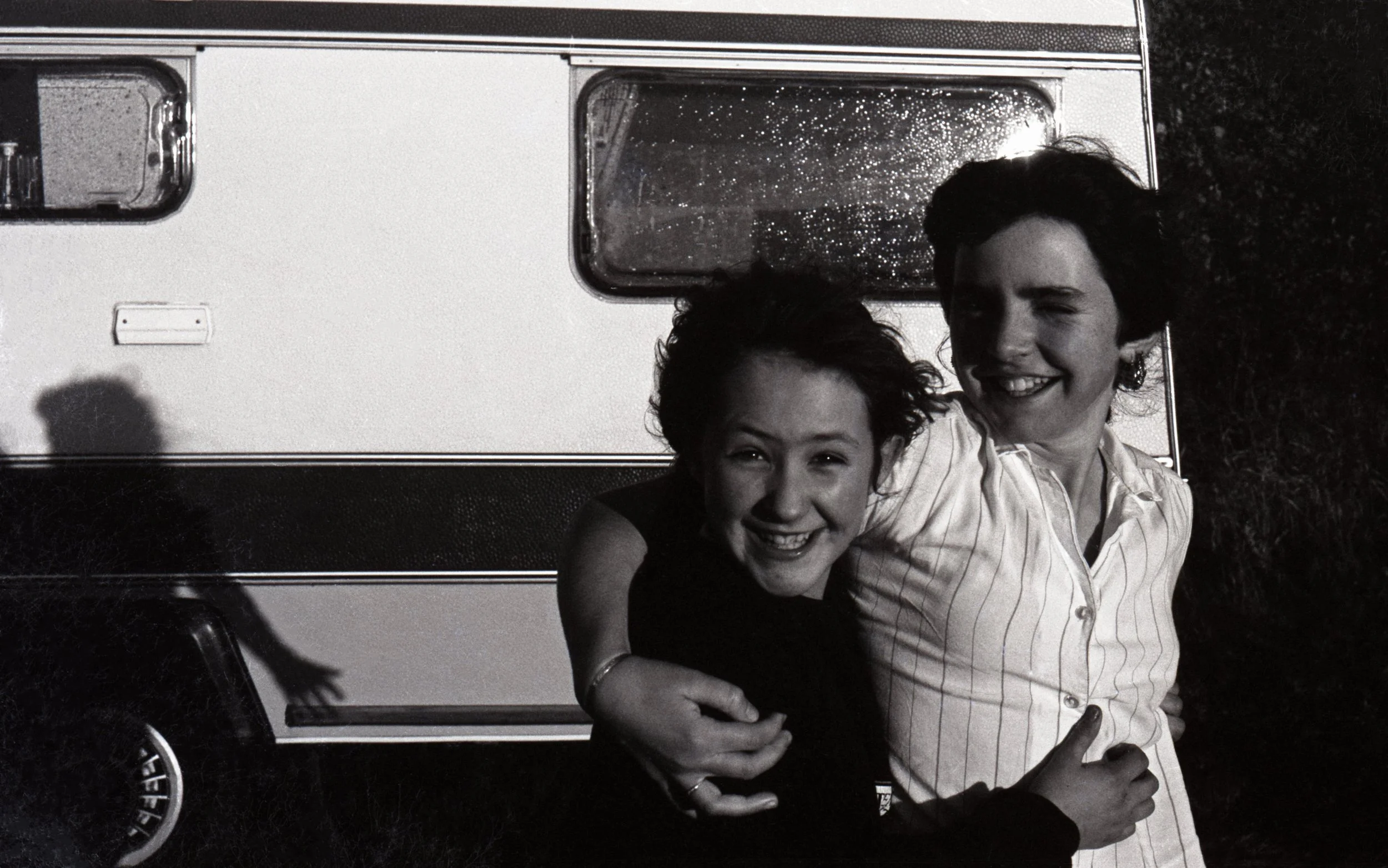 Black and white photo of two women hugging and smiling in front of a camper trailer, with one of them winking and making a peace sign.