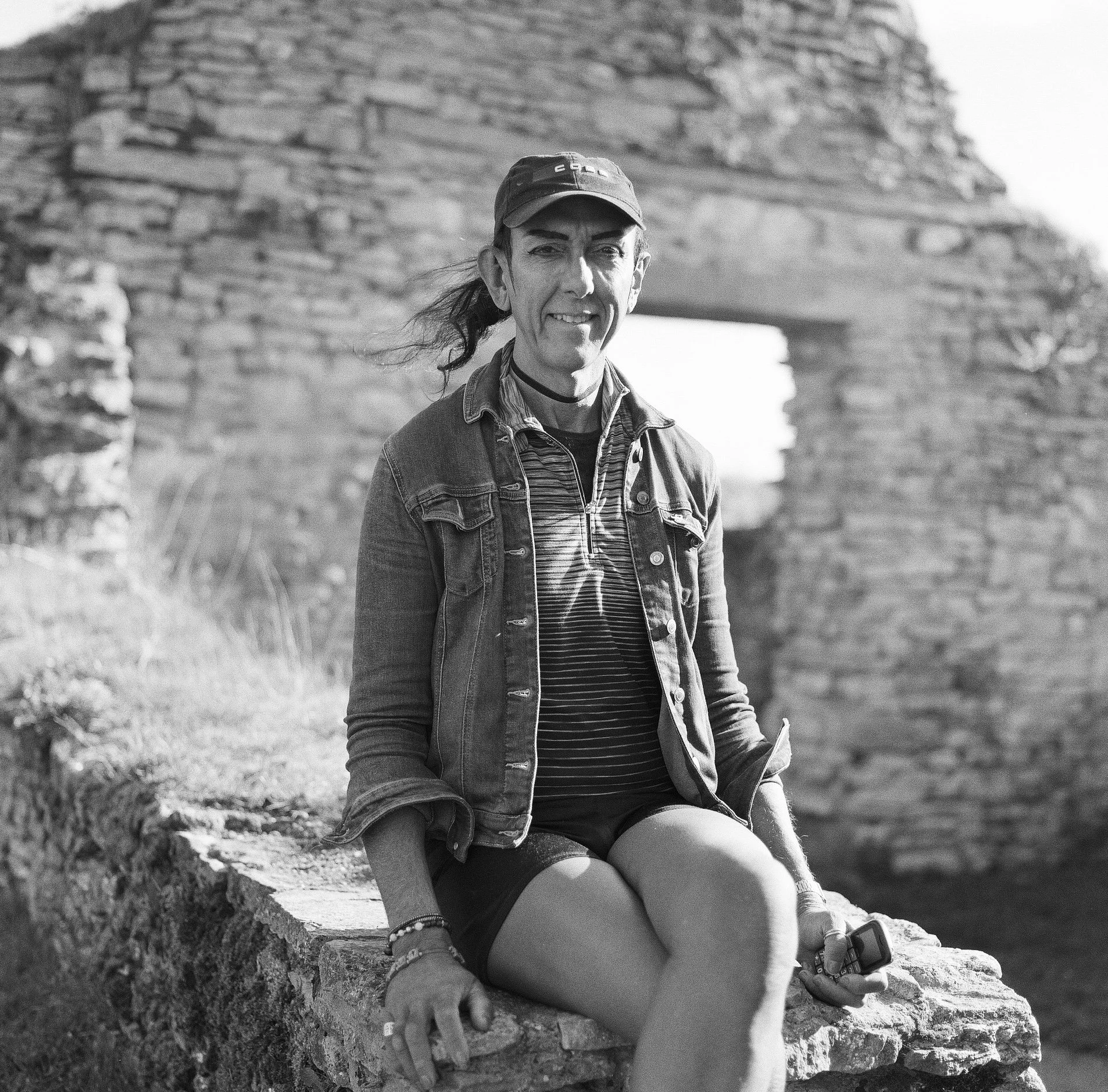 A woman sitting on a stone ledge outdoors, wearing a baseball cap, denim jacket, striped shirt, shorts, and sneakers, holding a smartphone with a scenic background.