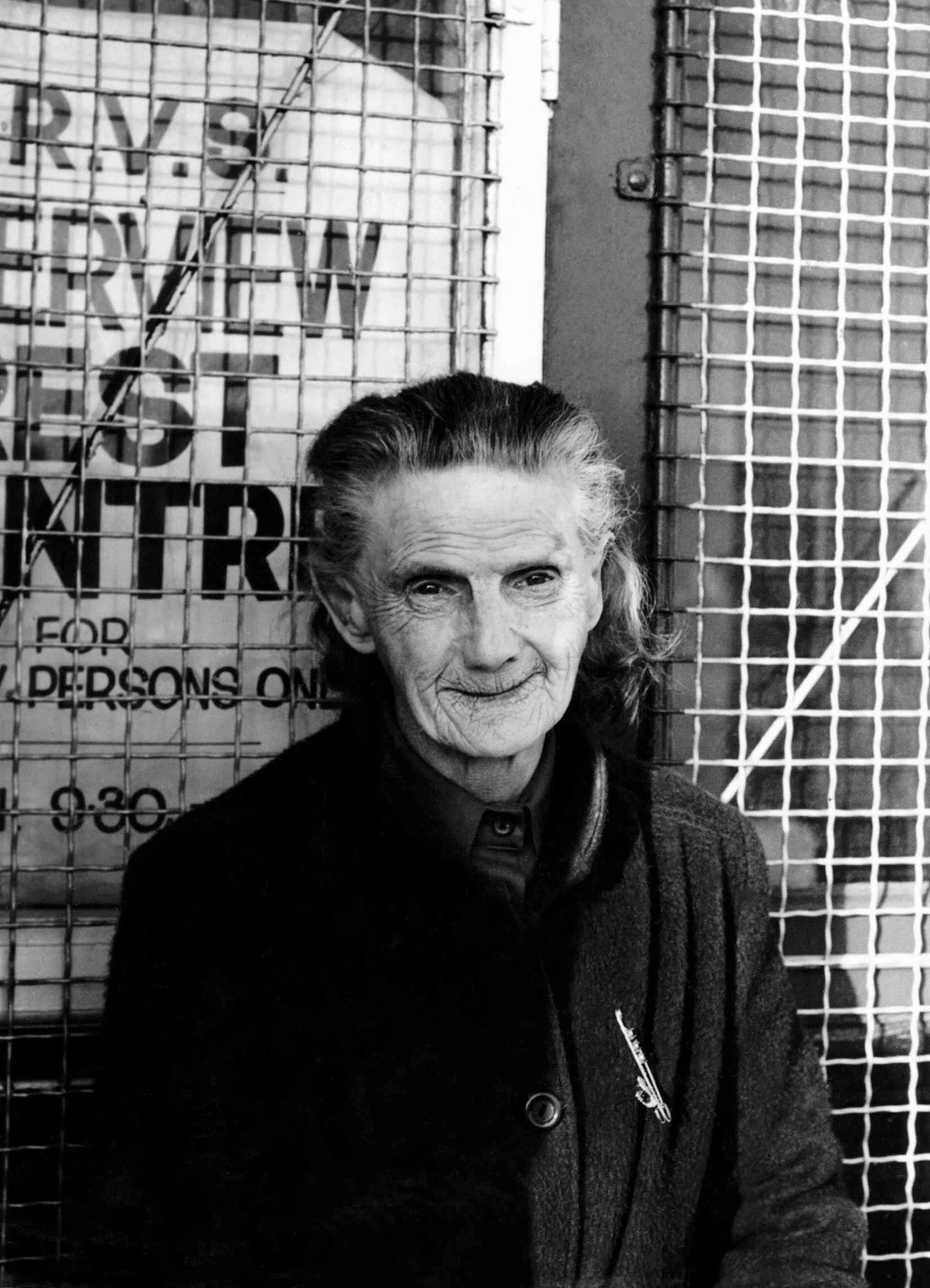A black and white photo of an elderly woman with smooth hair, smiling slightly, holding a pen, and standing in front of a metal grid background with warning signs.