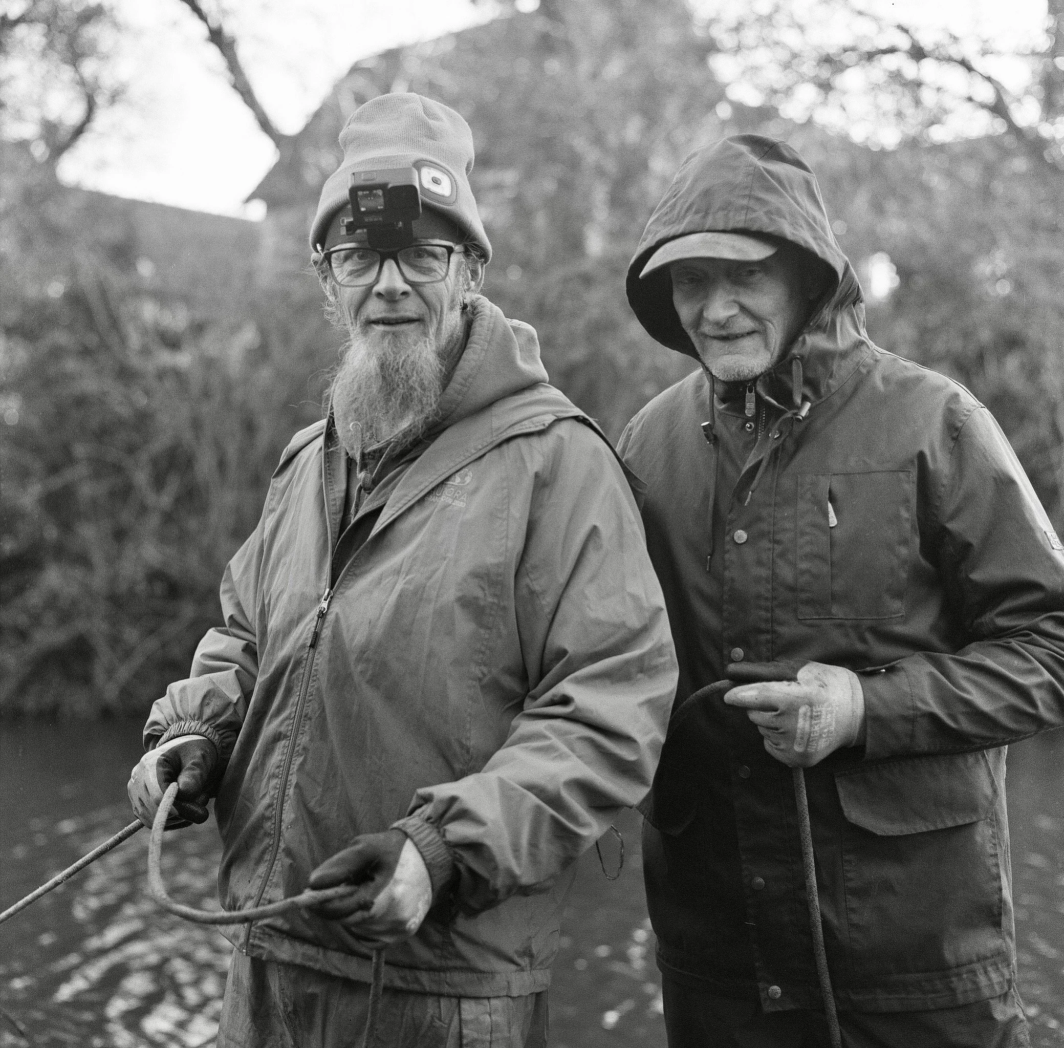 Two middle-aged men walking outdoors on a trail in a wooded area, with one wearing glasses, a beanie hat with a mounted camera, and a jacket, and the other wearing a hooded jacket with the hood up. They are holding a leash, and their expressions seem