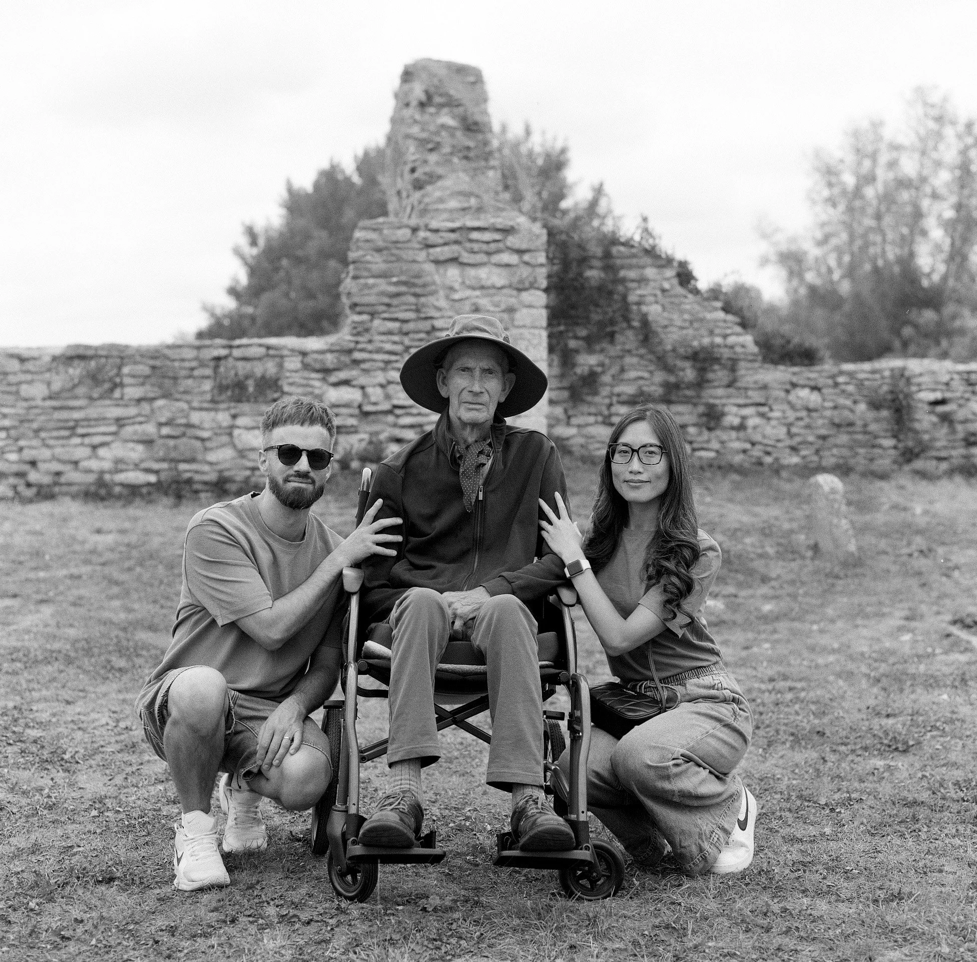 Four people outdoors in front of ancient stone ruins. Two women and two men, one man sitting in a wheelchair, the others standing close together, smiling, with trees in the background.