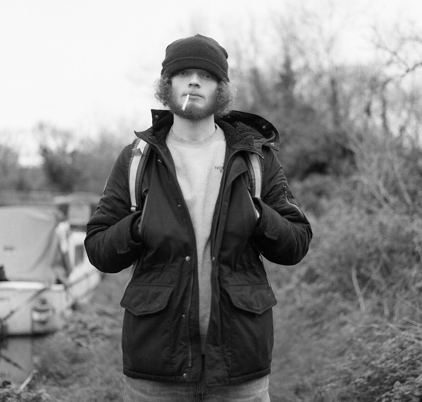 A young man with curly hair and a beard, wearing a beanie and a jacket, standing outdoors with trees in the background. He is holding a cigarette in his mouth.