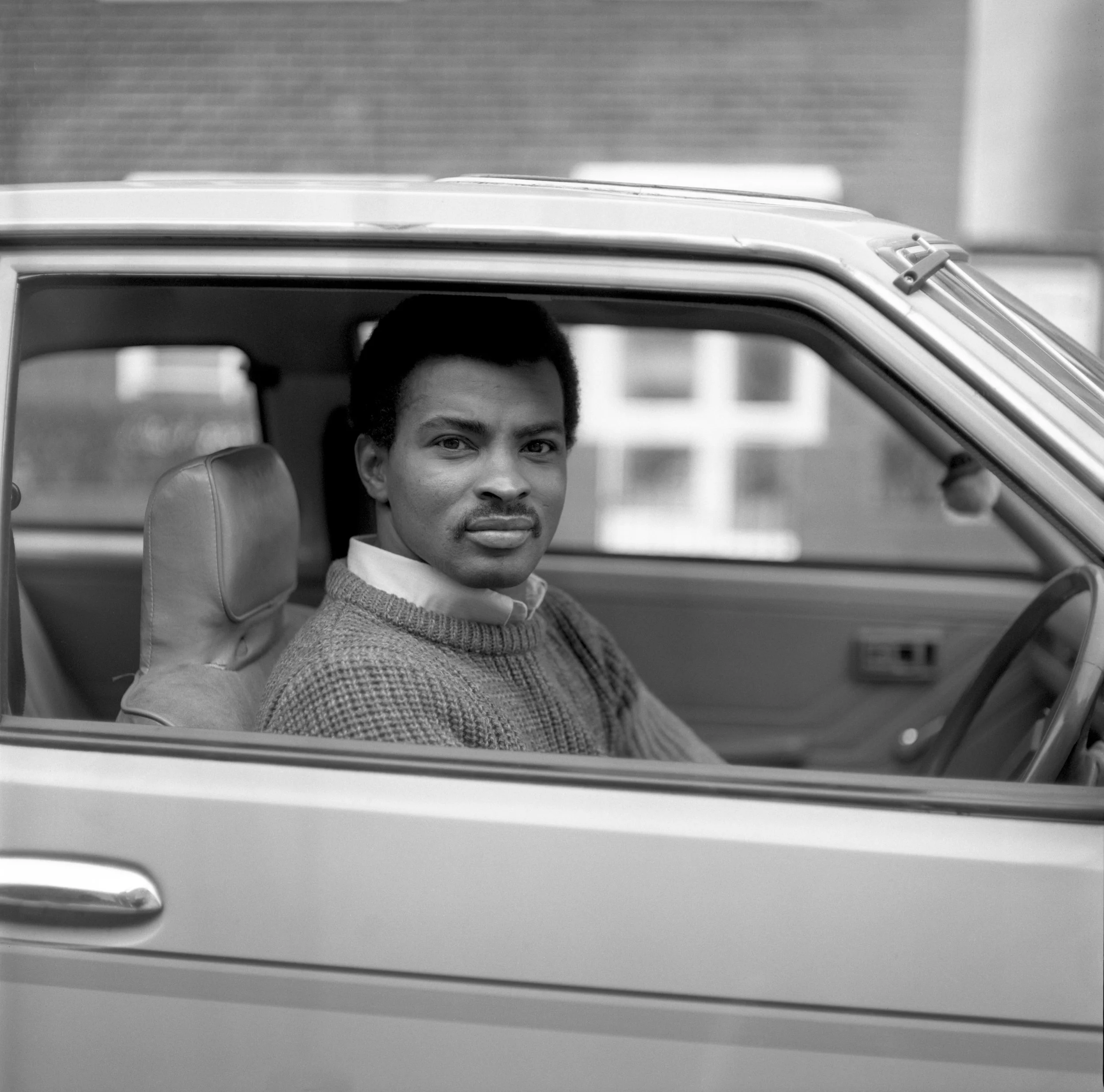 A black and white photo of a young man with dark hair and a mustache, sitting in a car, looking at the camera.