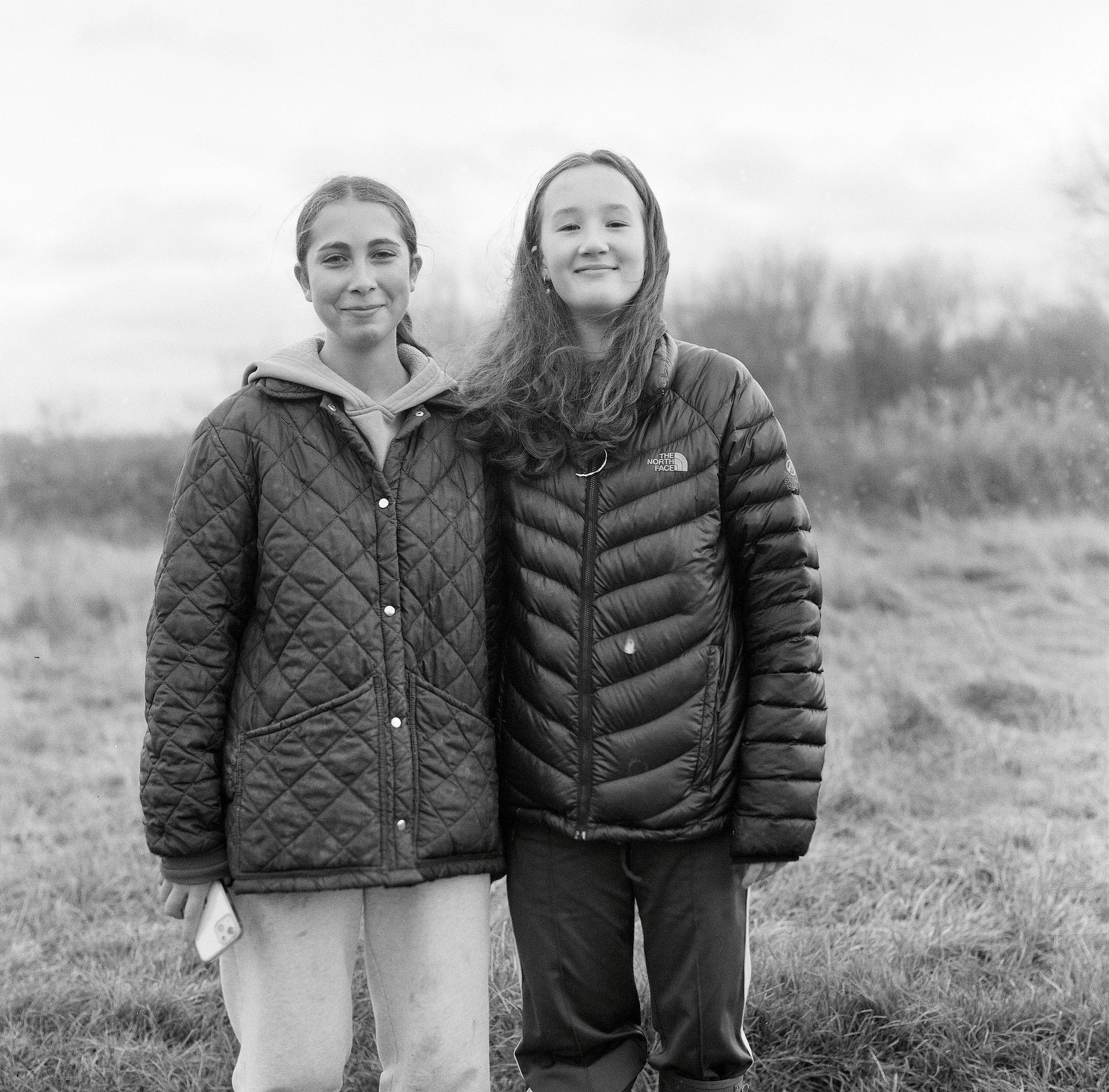 Two young girls standing in a grassy area outdoors, smiling at the camera, with trees and cloudy sky in the background.