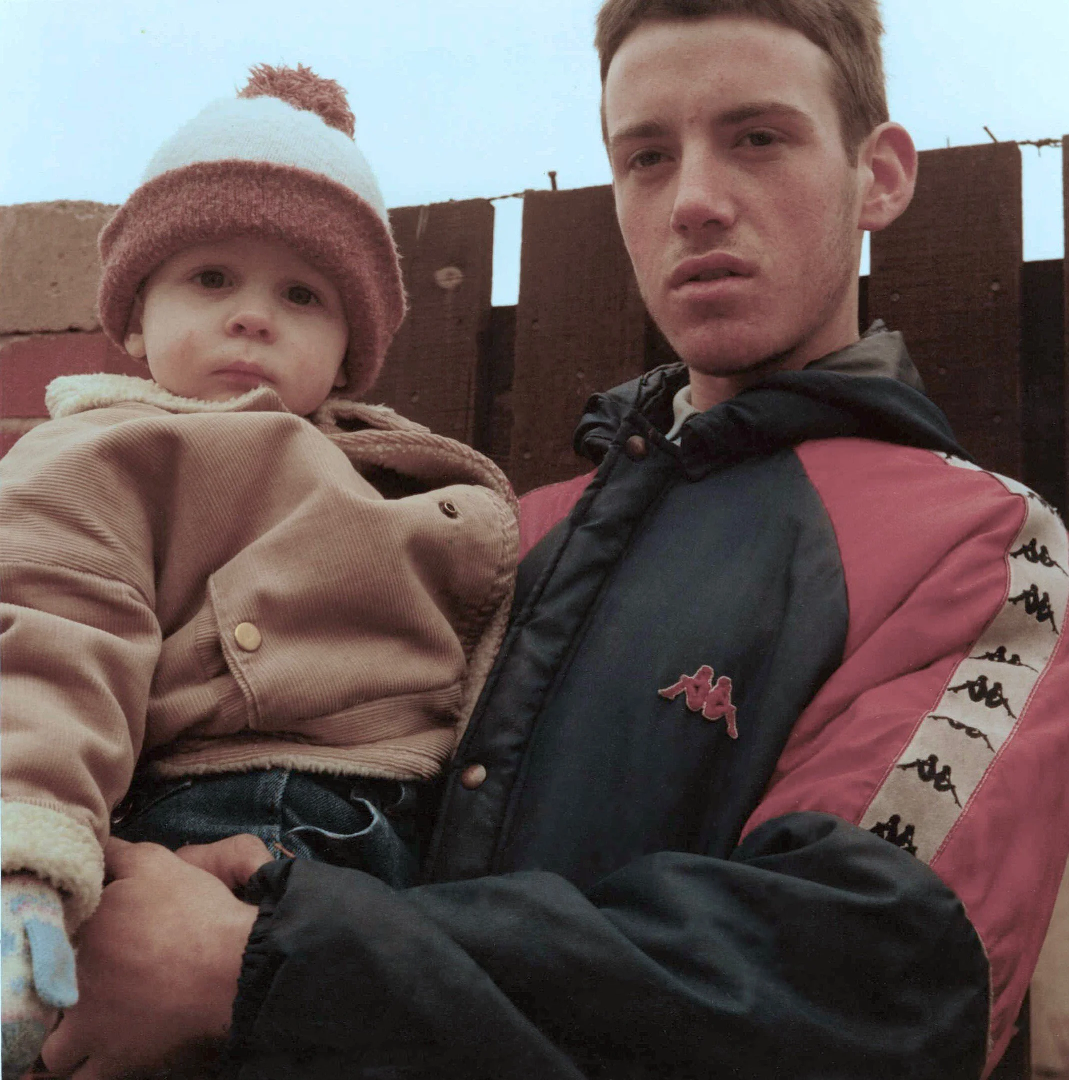A young man with short red hair holding a small child with light hair and a pink and white knit hat, against a background of a brown wooden fence and cloudy sky.