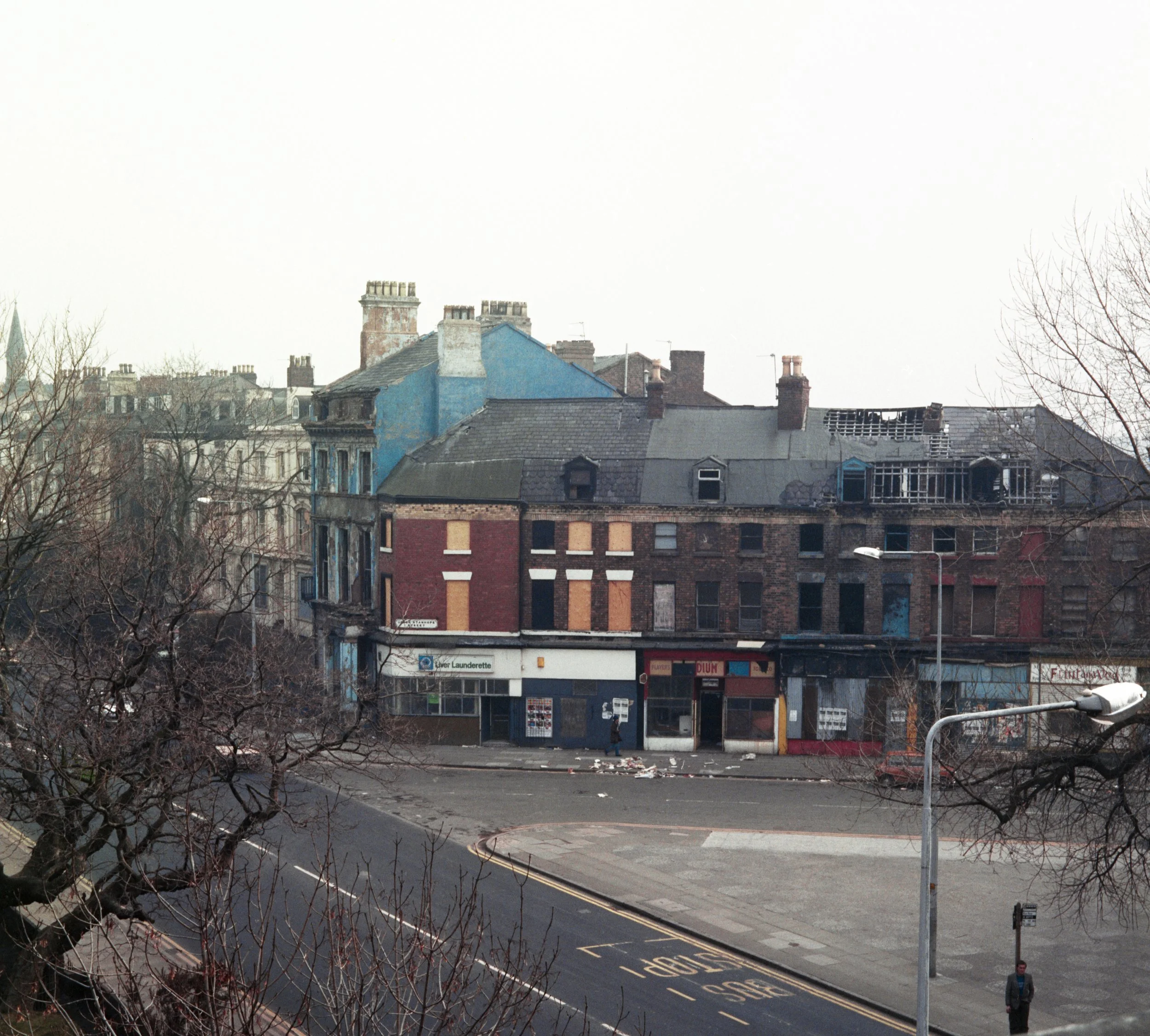 Cityscape with rooftops, chimneys, leafless trees, and a parking lot with some people and cars visible in an urban area.