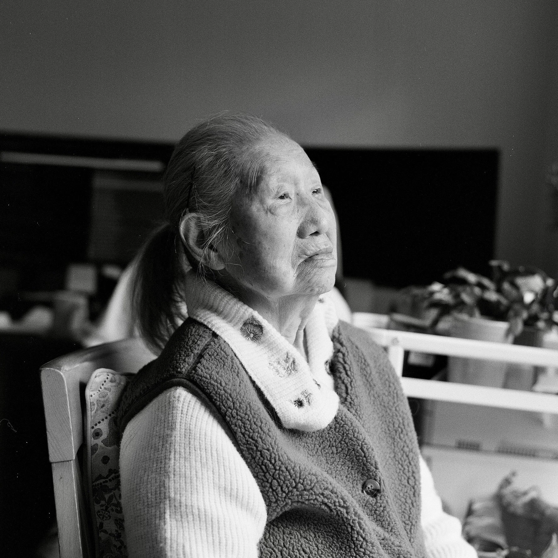 A black and white photo of an elderly Asian woman sitting on a chair, looking slightly to the side with a thoughtful expression, wearing a cozy sweater with a floral design and a fleece vest.