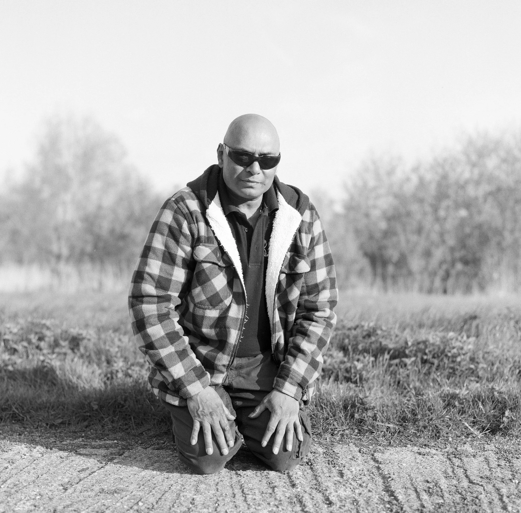 A man in sunglasses wearing a checkered jacket kneels on the ground with one hand on the ground and the other on his knee outdoors in a field.