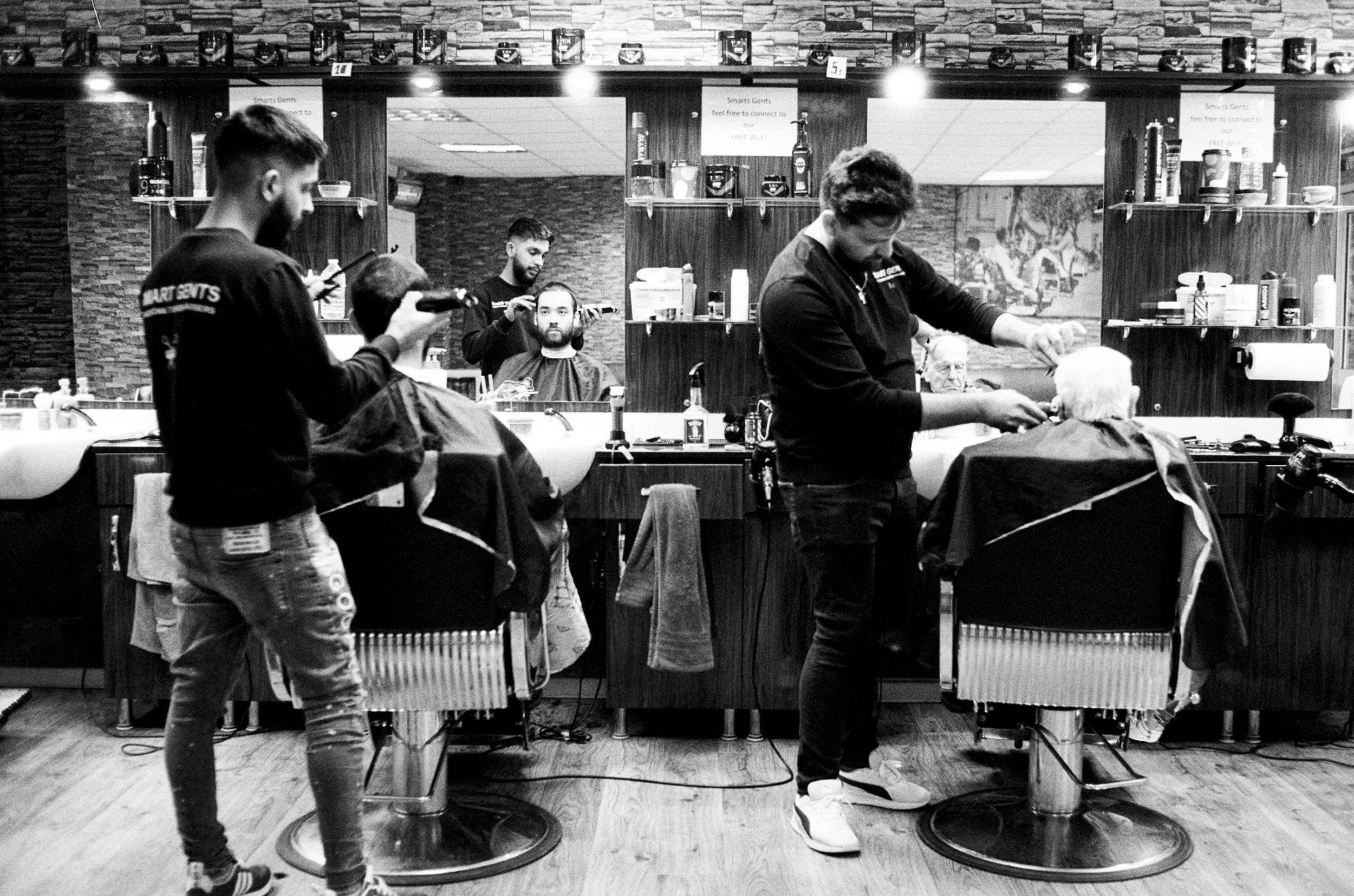 Barbershop scene with three barbers cutting clients' hair and a customer waiting. Barbers are focused on their work, and the shop has shelves with hair product bottles, mirrors, and a rustic brick wall background.