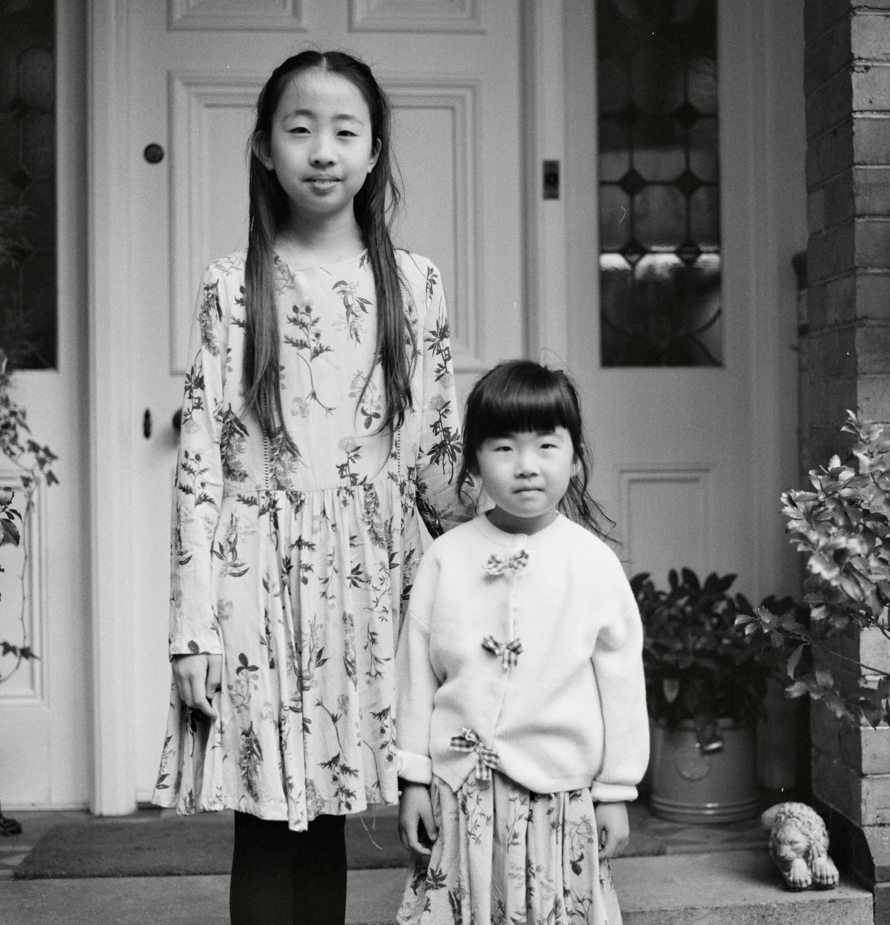 A black and white photo of two young girls standing outside in front of a house. The older girl is wearing a long floral dress and has long hair; she is smiling slightly and has her arm around the younger girl. The younger girl is wearing a light-col