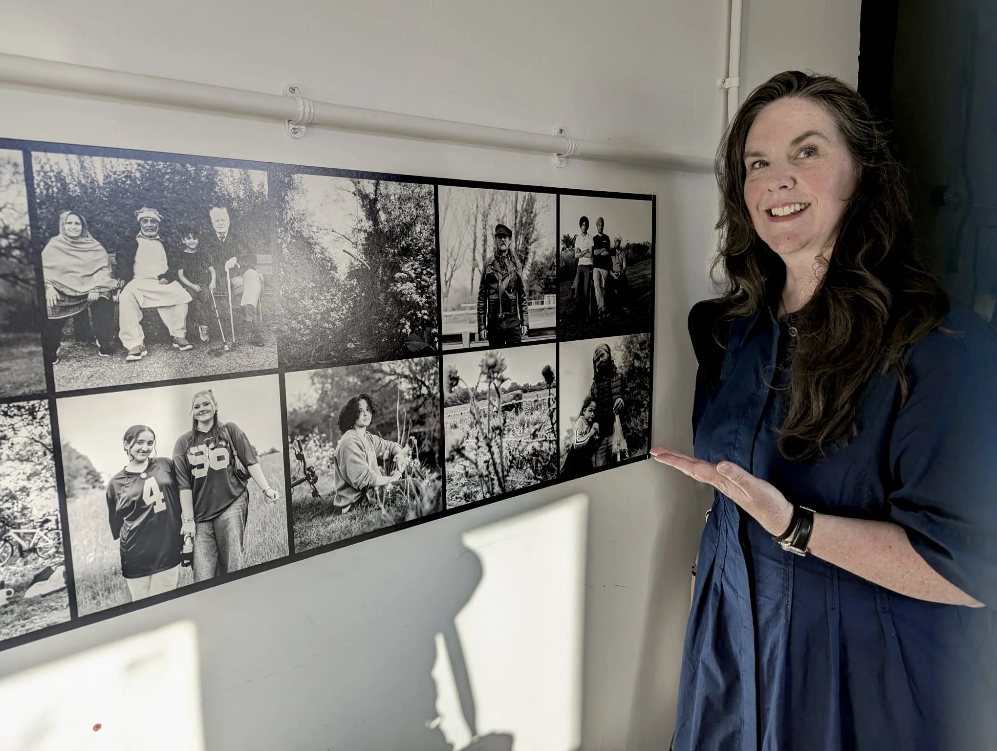 A woman with long dark hair smiling and pointing at a collage of black-and-white photographs on a wall.