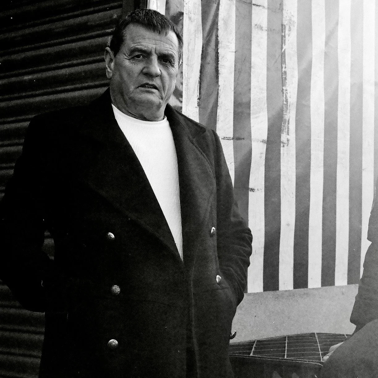 A black and white photo of an older man with short dark hair, wearing a double-breasted coat and a light-colored shirt, standing next to a wall with vertical stripes.