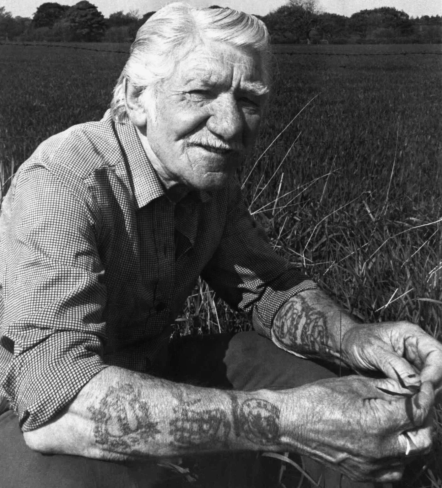An elderly man with white hair and a mustache, sitting outdoors in a field of tall grass, smiling and looking at the camera.