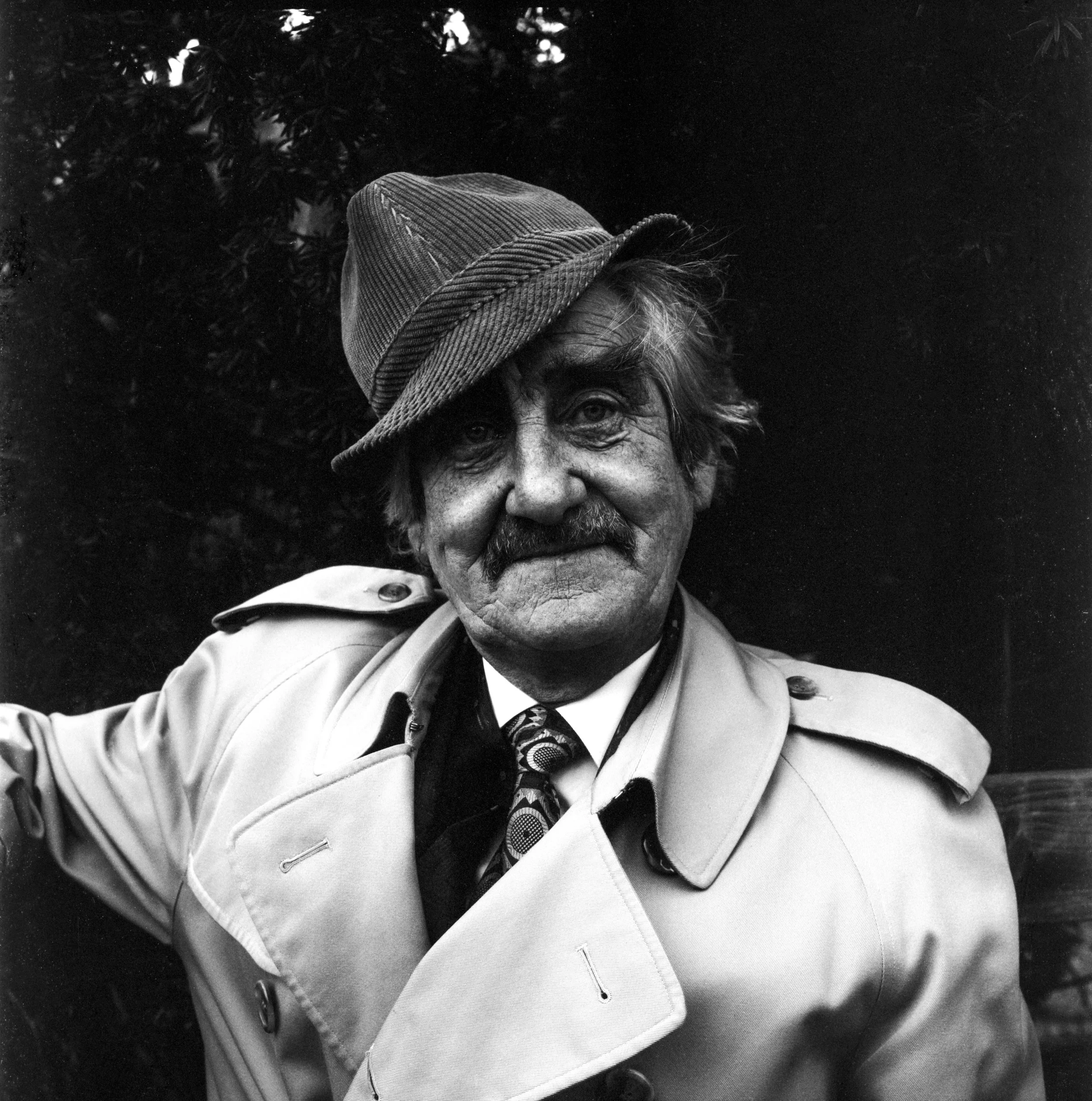 A black and white photograph of an elderly man wearing a fedora hat, a trench coat, and a patterned tie, with trees in the background.