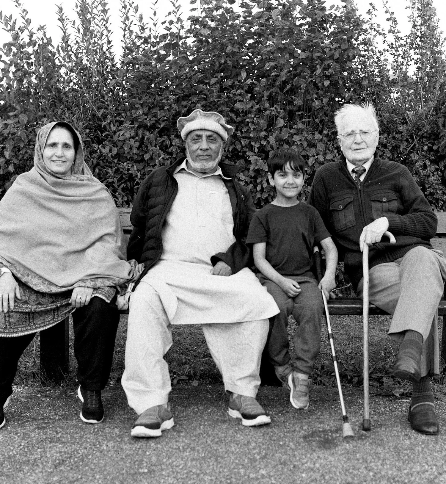 Four people sitting on a park bench in front of leafy bushes, two adult men, a woman, and a young boy. The woman is wrapped in a shawl, and the man in the center is wearing a hat. The man on the right has white hair, glasses, and a cane.