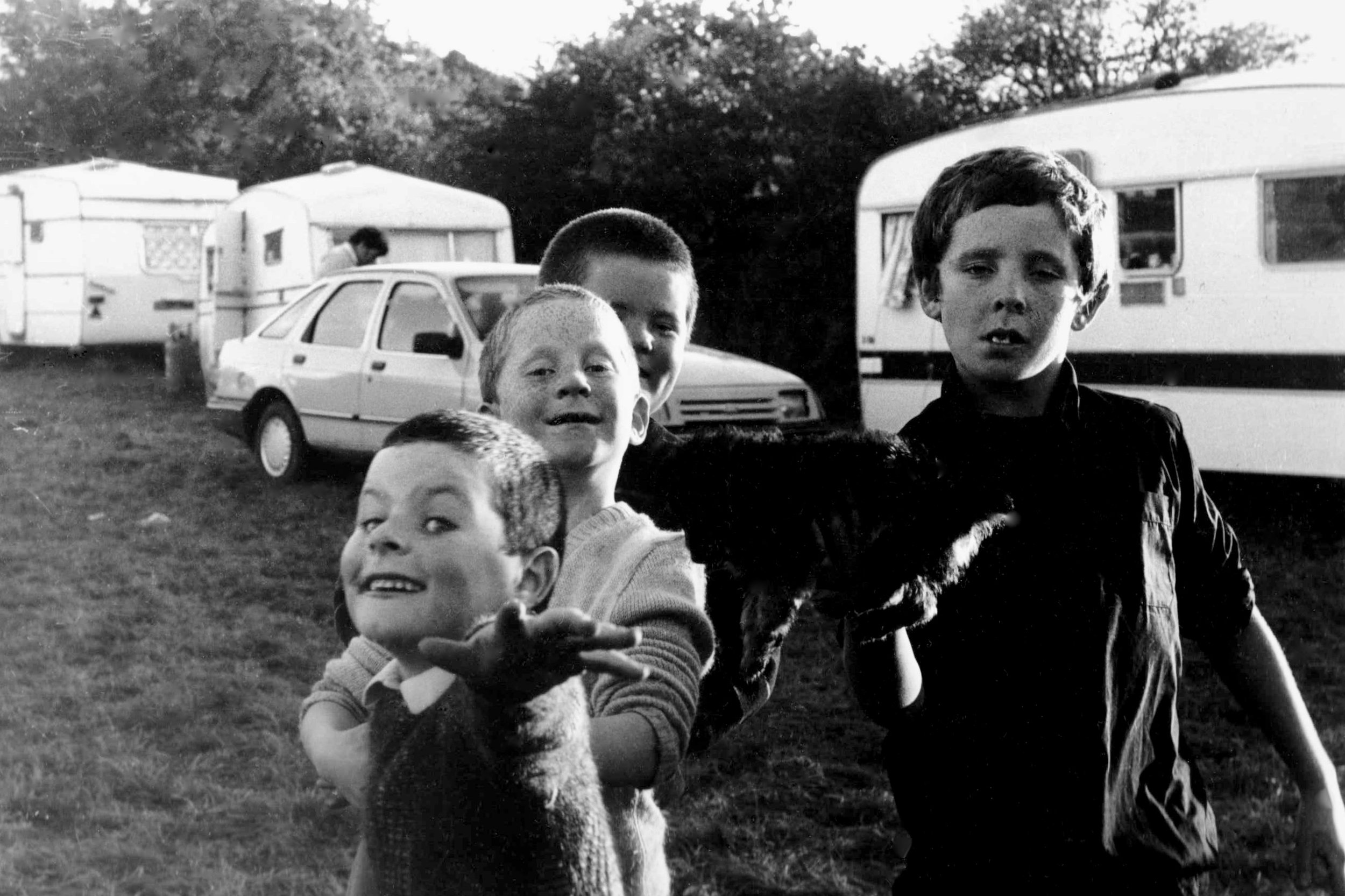 Black-and-white photo of several children playing outdoors near parked cars and caravans, with trees in the background.