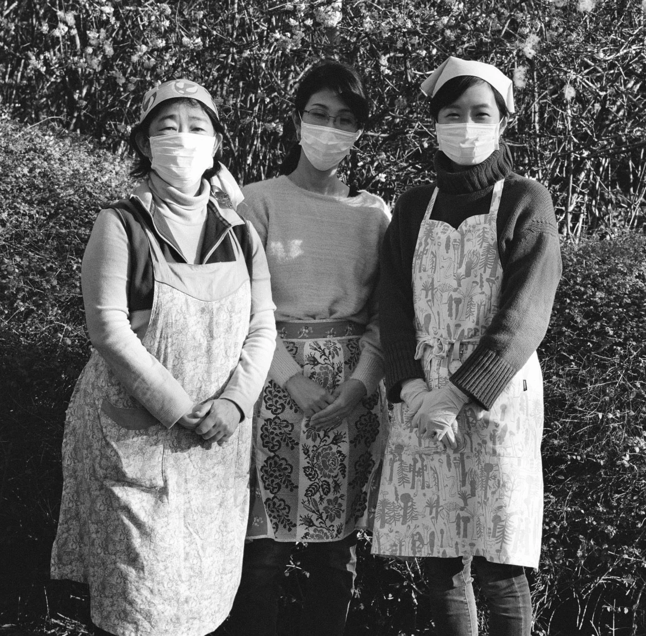 Three women wearing masks and aprons standing outdoors in front of bushes.