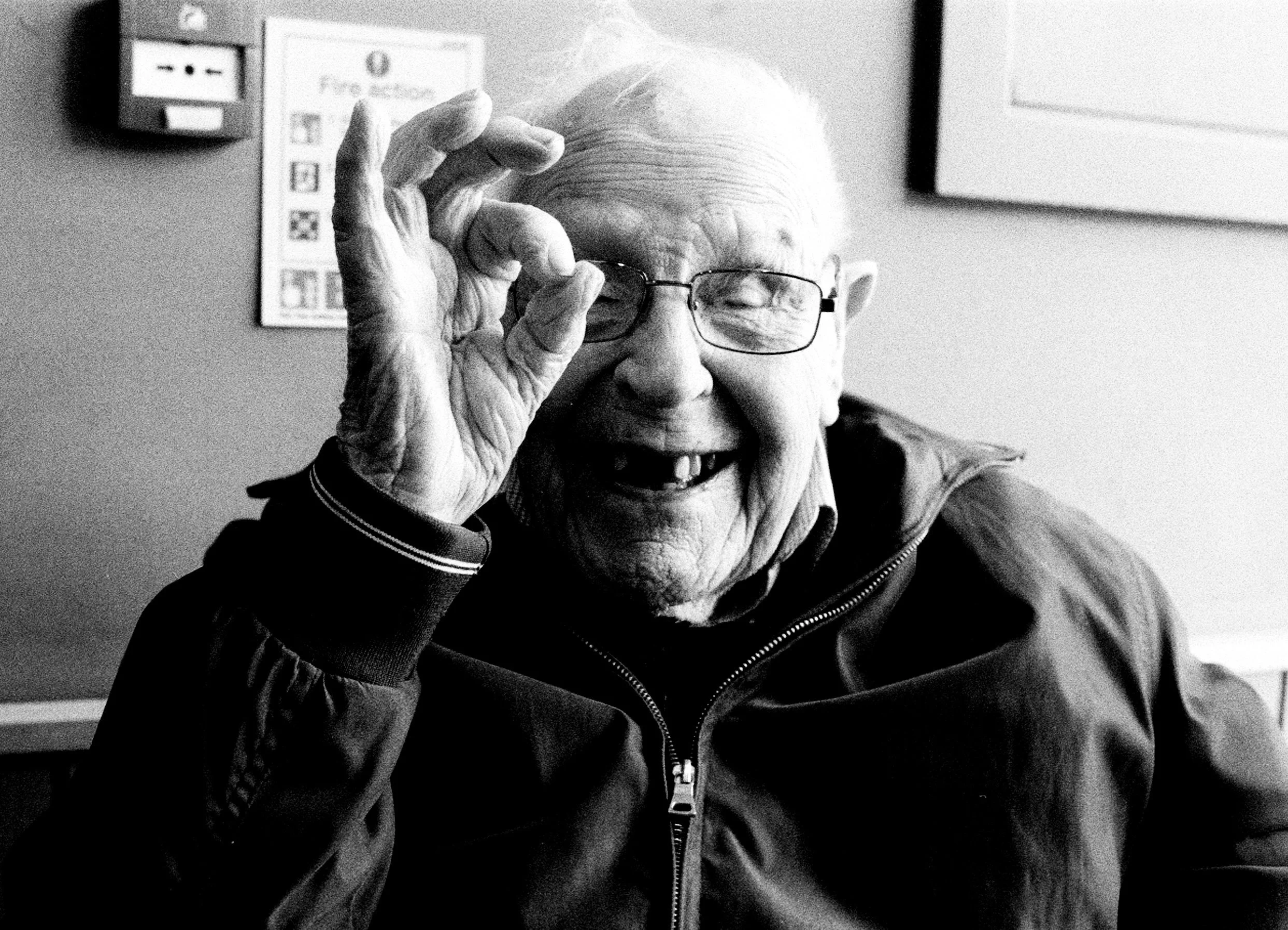 An elderly man with glasses smiling and holding his glasses with his hand. He is wearing a jacket and is in an indoor setting with posters or signs on the wall in the background.
