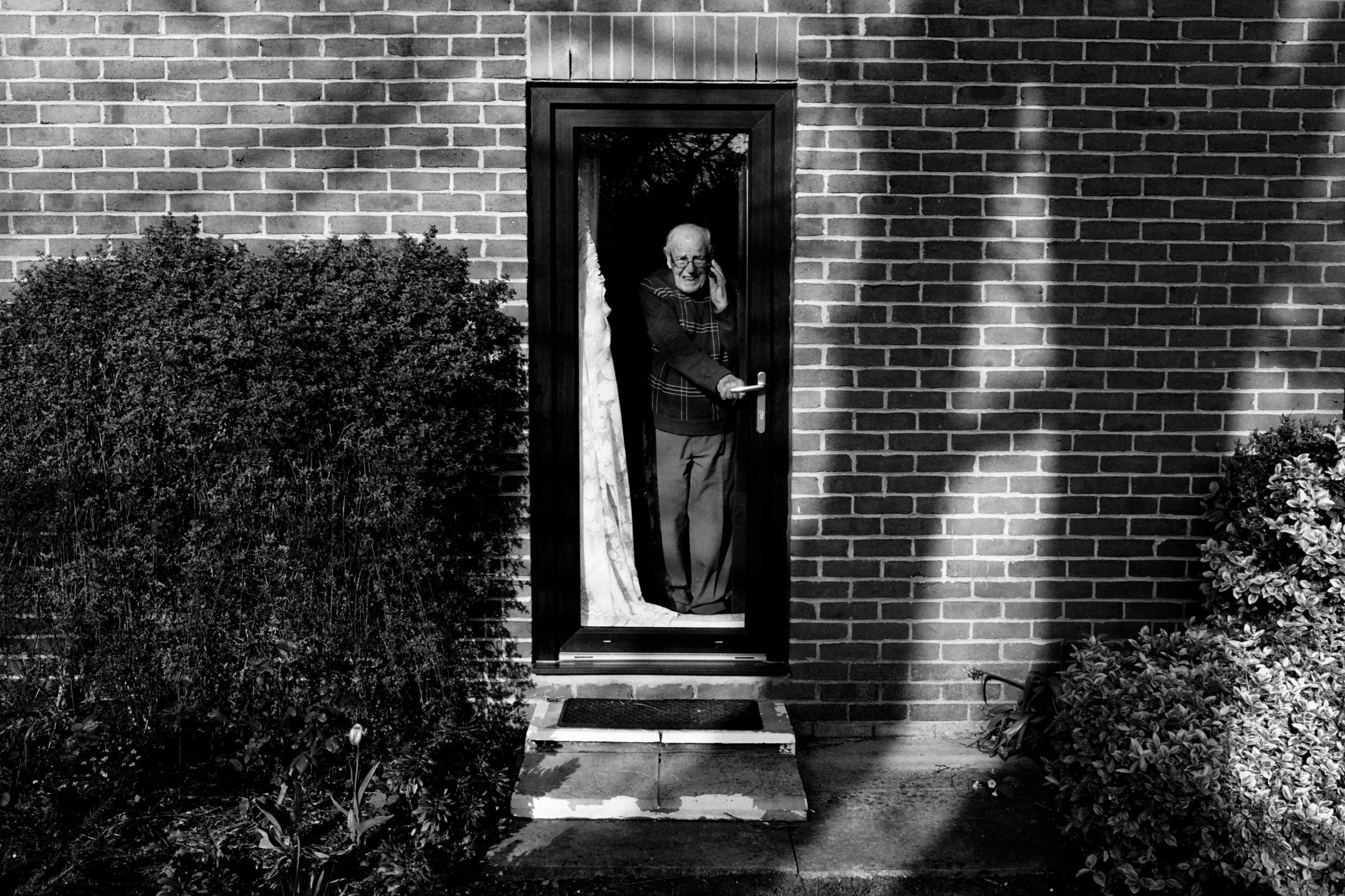 An elderly woman with glasses smiling and talking on a cell phone while standing in a small glass porch outside a brick house at night. She is smiling and holding the door handle with her other hand.