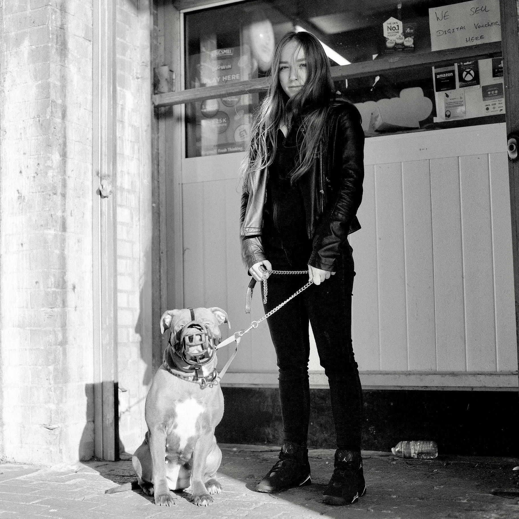 A young woman in dark clothing and sneakers stands outside a shop, holding a leash attached to a dog. The dog, possibly a Pit Bull, sits on the sidewalk, looking up at her. The shop window behind them has signs and items displayed, including a sign t
