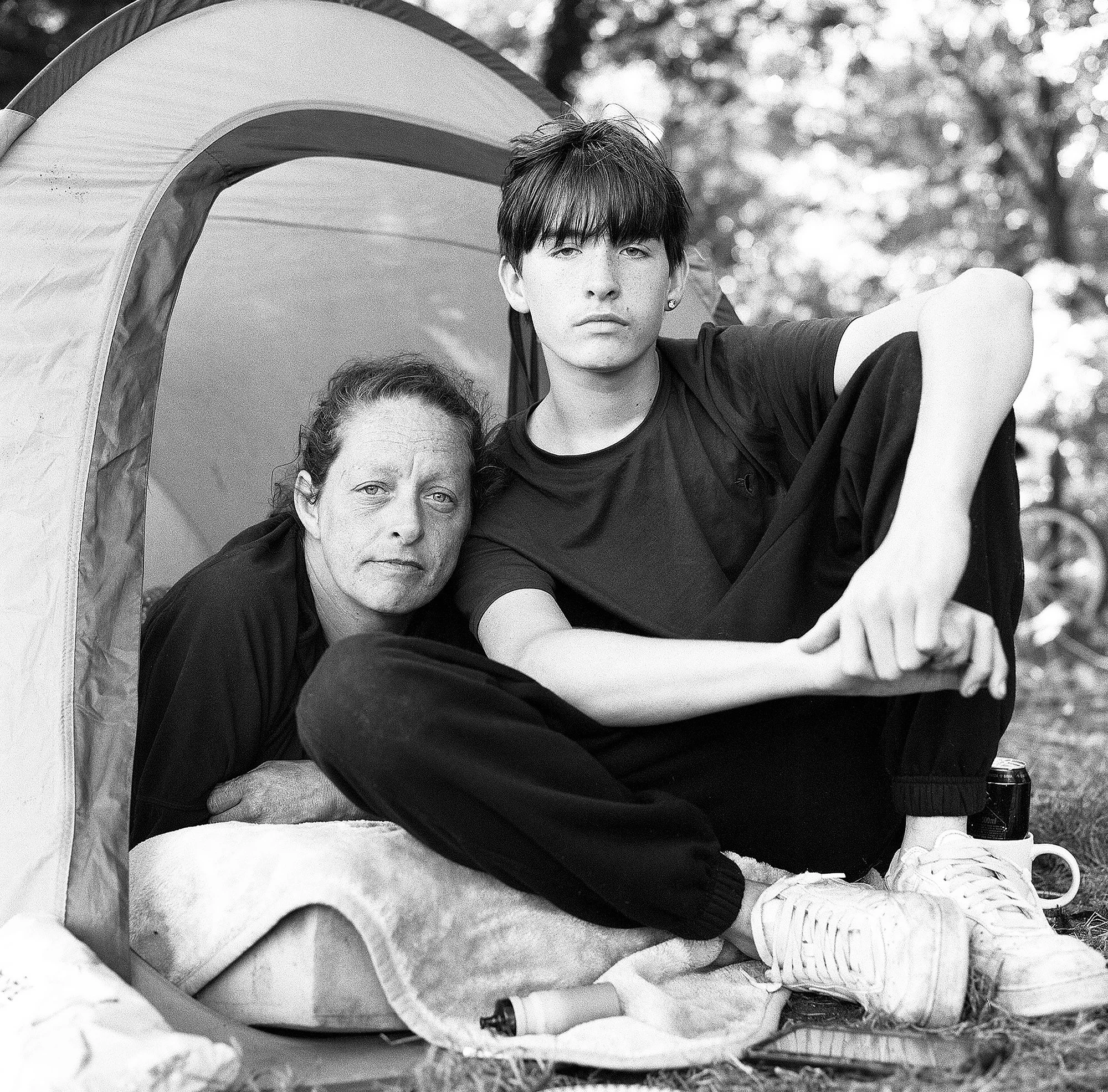 Black and white photo of a young man with dark hair, wearing a black t-shirt, leaning on a sleeping woman with outstretched arm, under a curved structure, outdoors with blurred trees in the background.