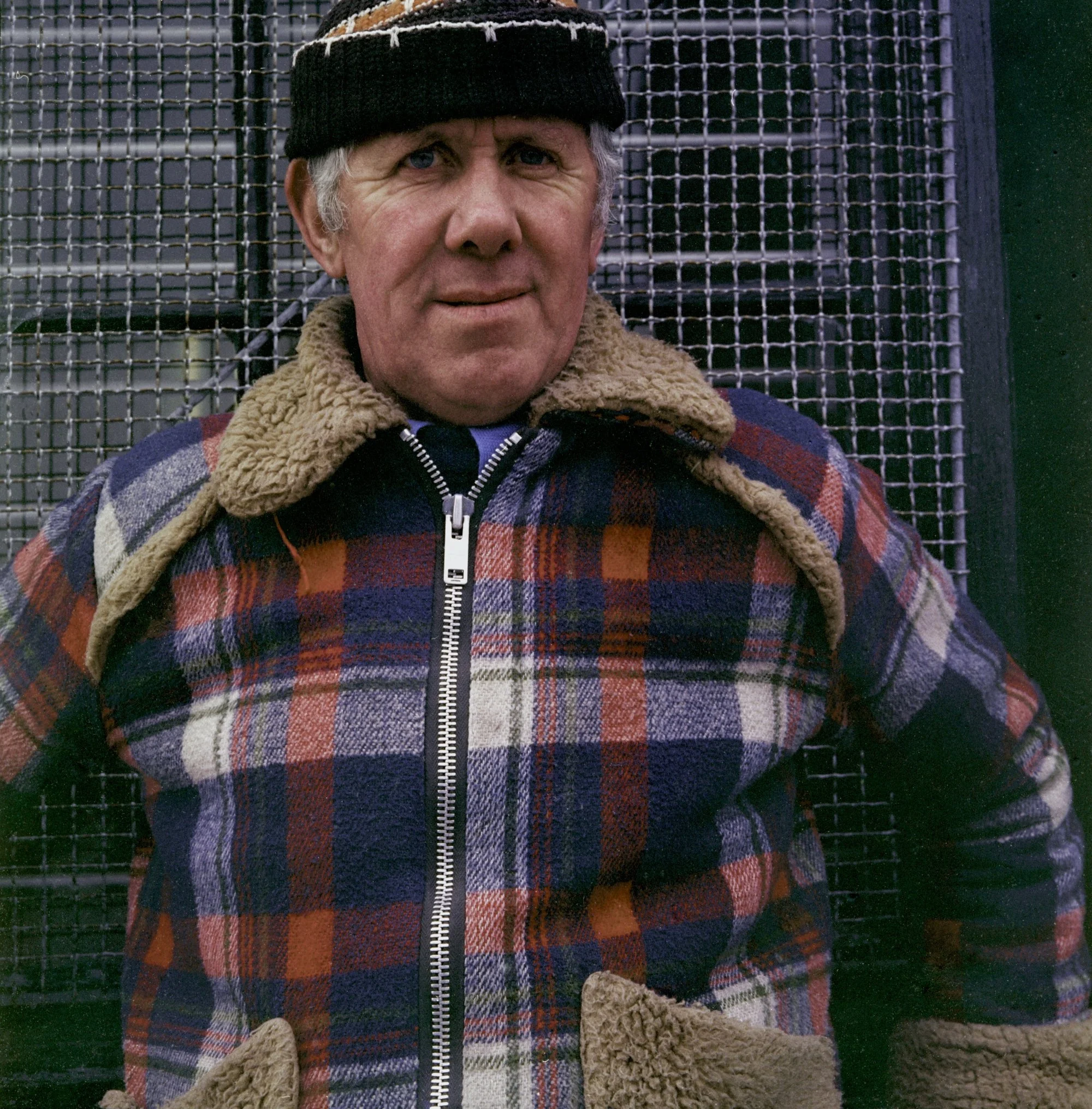 A man with gray hair and blue eyes wearing a black knit cap, a plaid coat with a zip, and a brown fleece-lined jacket sitting in front of a metal fence.