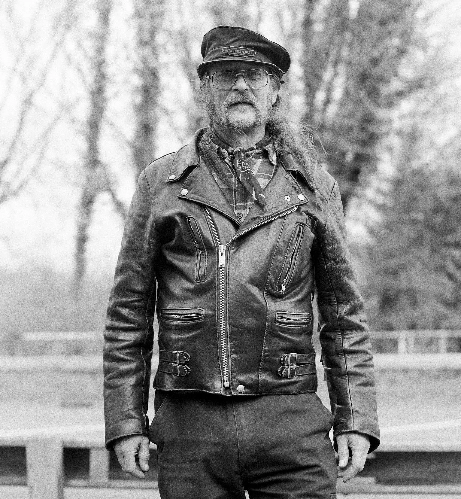 A black-and-white photo of a man with long hair, a beard, and glasses, wearing a leather biker jacket, a plaid shirt, a bandana around his neck, and a cap with the inscription 'British Railways.' He is outdoors with blurred trees in the background.
