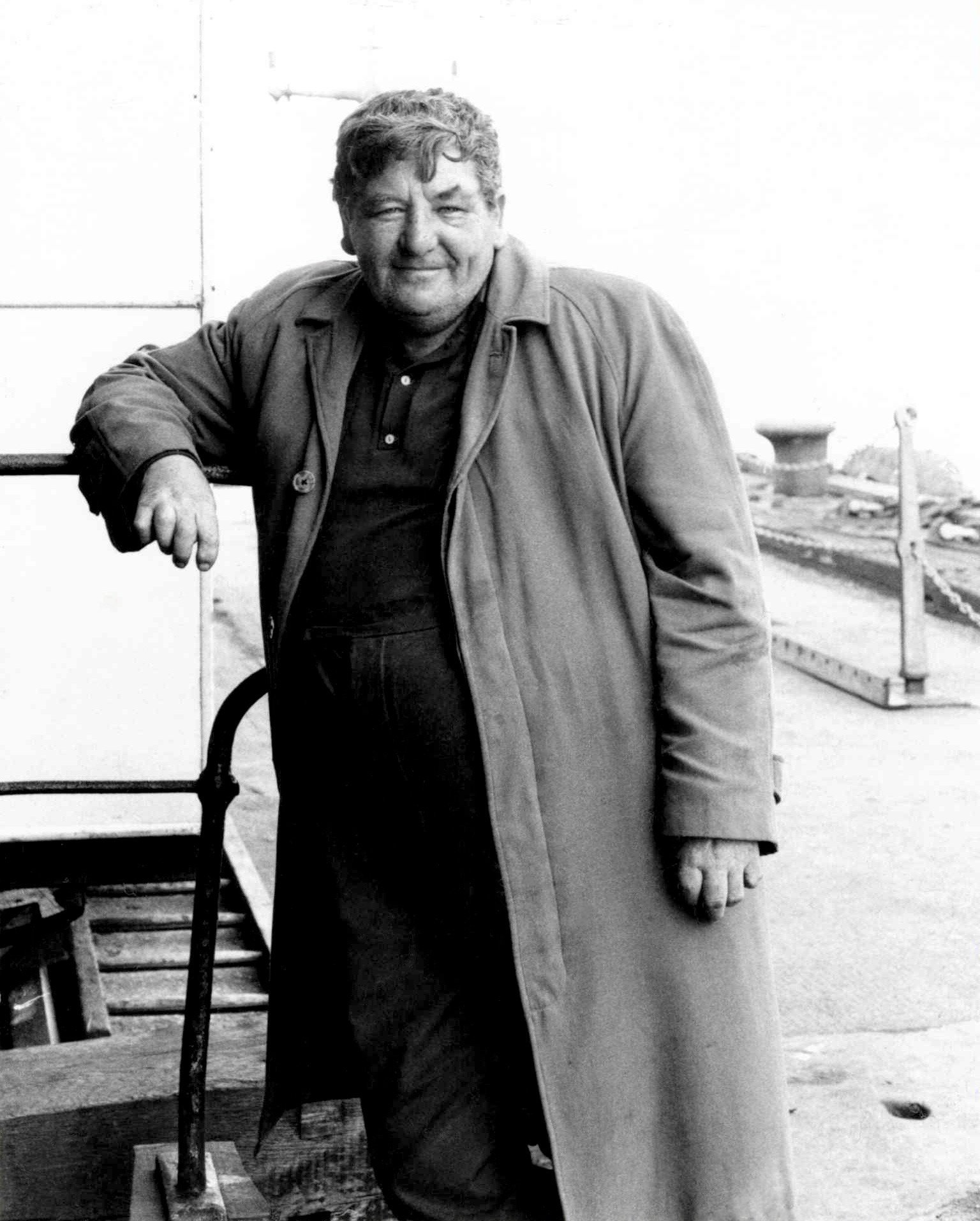Black and white photo of a man with curly hair leaning on a wooden chair outdoors, with a blurred background of a fence and a flat landscape.