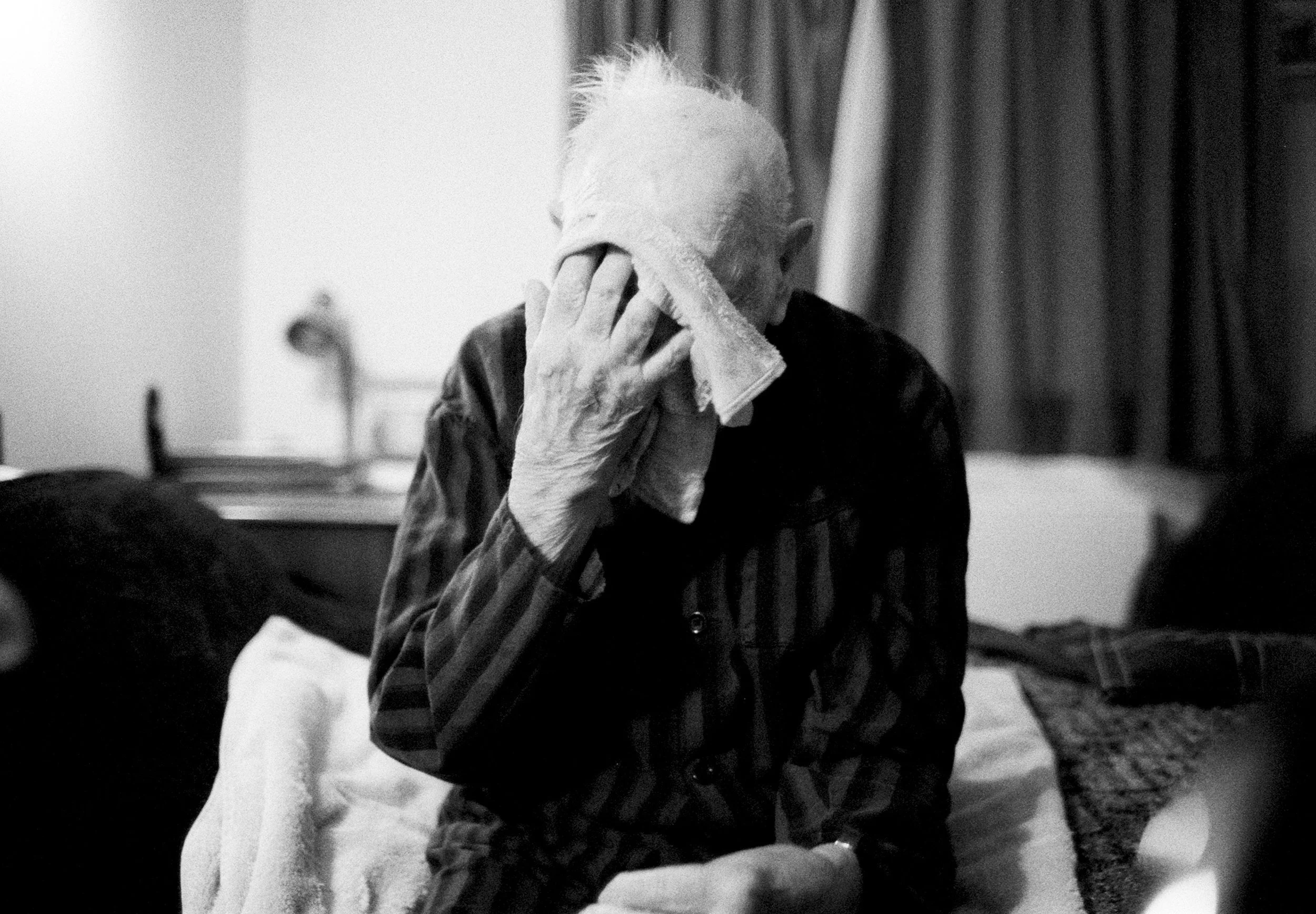An elderly person with white hair, dressed in a dark plaid shirt, wiping their face with a towel. The background shows dark curtains and a blurred room.