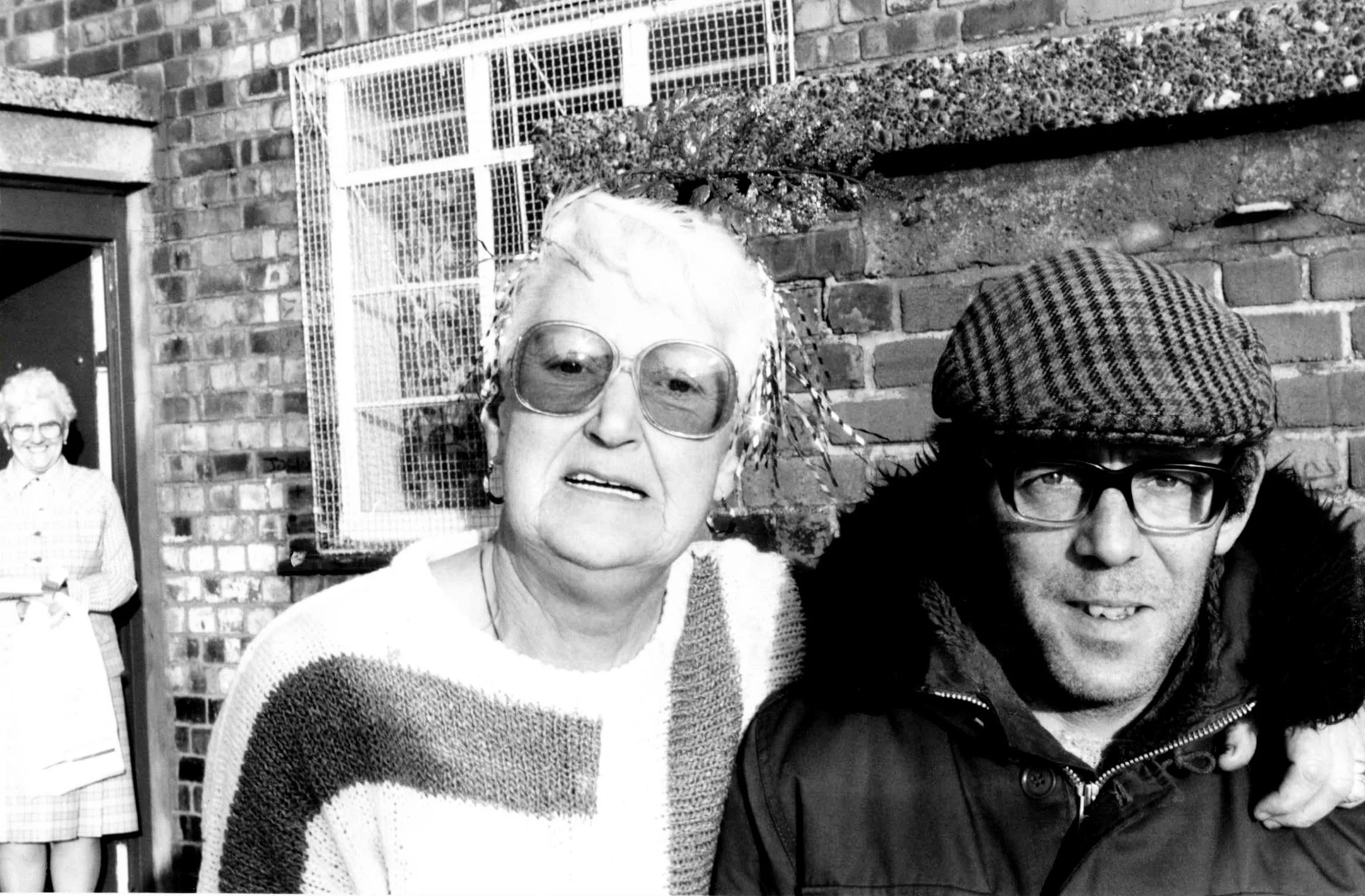 Black and white photo of two women in the foreground, one with glasses and short hair wearing a striped sweater, the other wearing large sunglasses and a jacket. In the background, a man with glasses and curly hair, and a girl in a checkered dress ar
