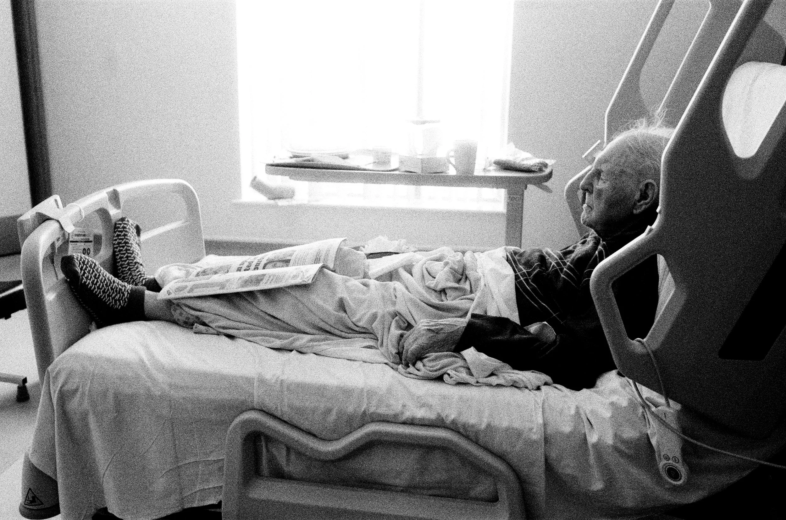 An elderly man with white hair lying in a hospital bed, wearing a checkered shirt and patterned socks, with a newspaper on his lap. The hospital room has a window with light streaming in, and medical equipment in the background.