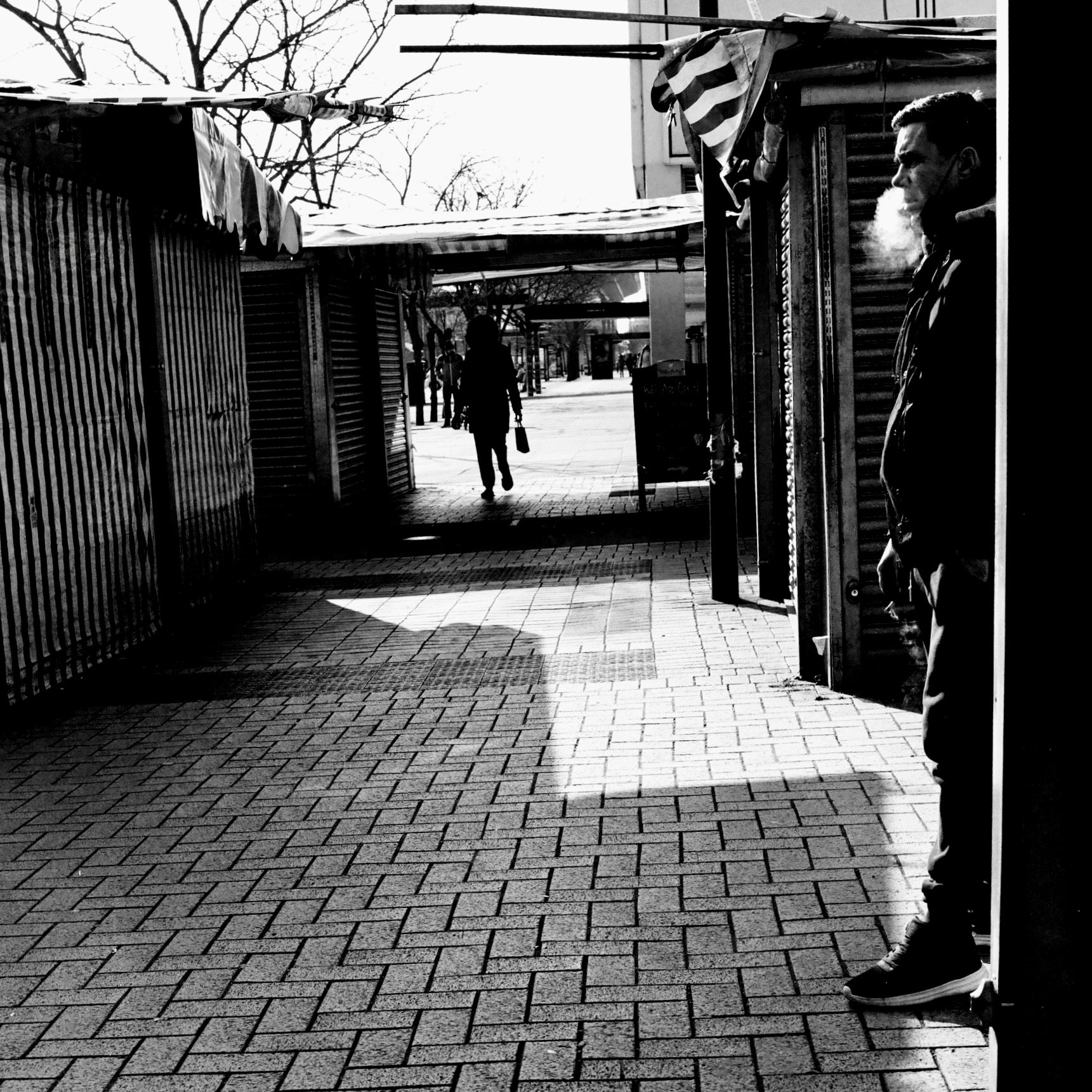 A black and white photo of a street scene with a person smoking outside a storefront on the right side. The person is wearing a jacket and sneakers, with visible breath vapor. In the background, a woman is walking away carrying a bag, and leafless tr