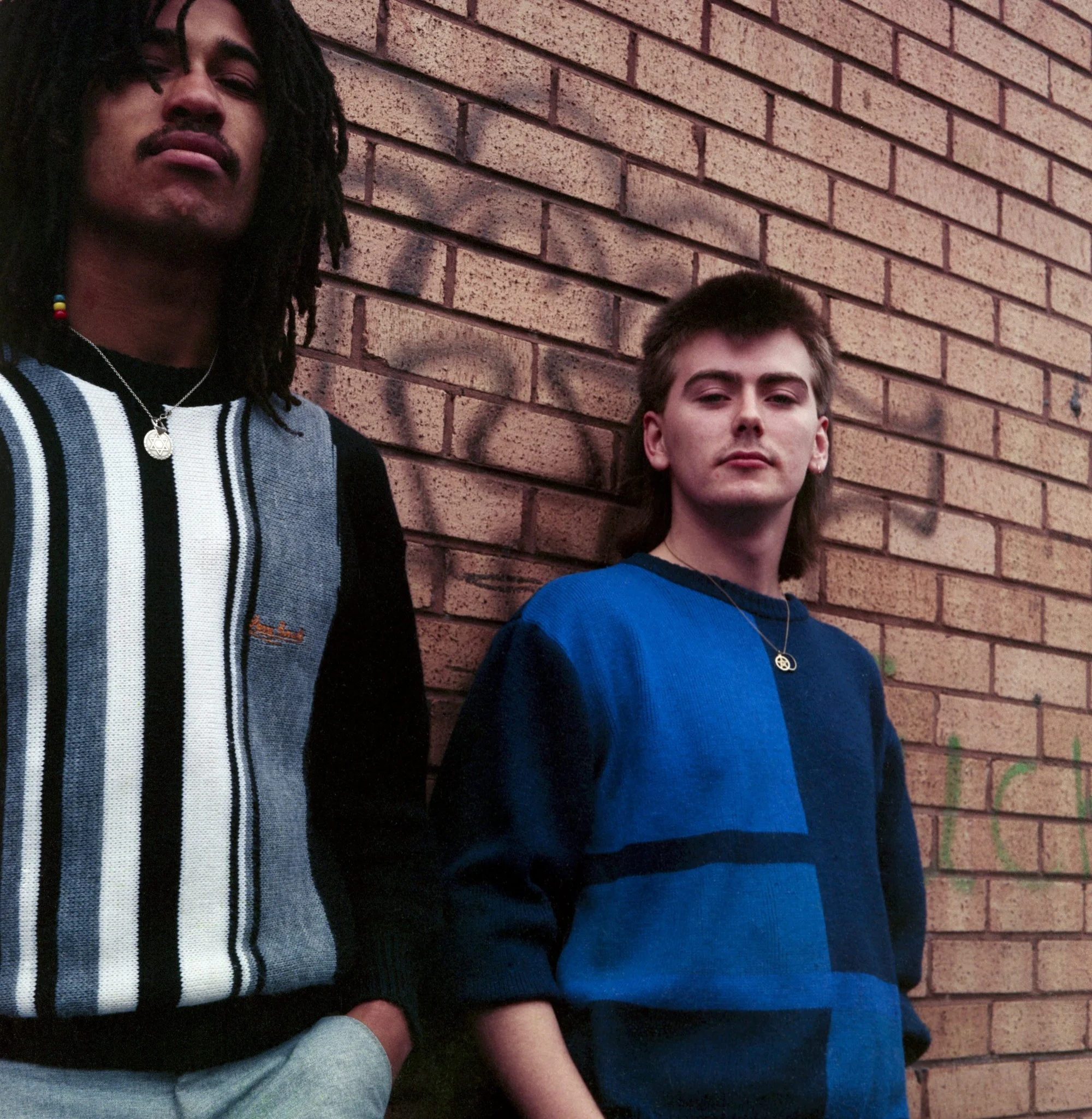 Close-up of two people with a brick wall background. One person has long black dreadlocked hair, wearing a striped sweater with a crescent moon pendant necklace. The other person has short light brown hair, wearing a blue t-shirt and a heart-shaped n