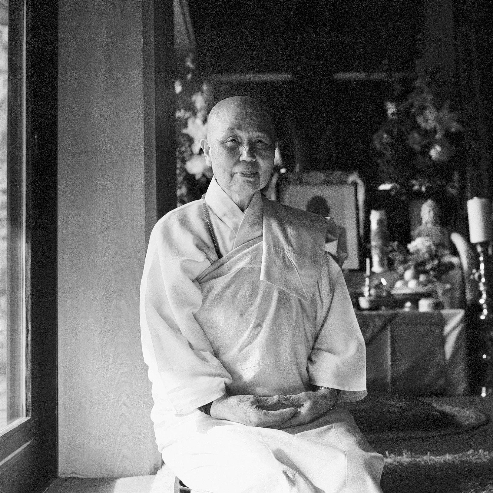 An elderly person with a shaved head wearing traditional clothing, sitting with hands clasped, smiling softly, in a room decorated with flowers and artwork.
