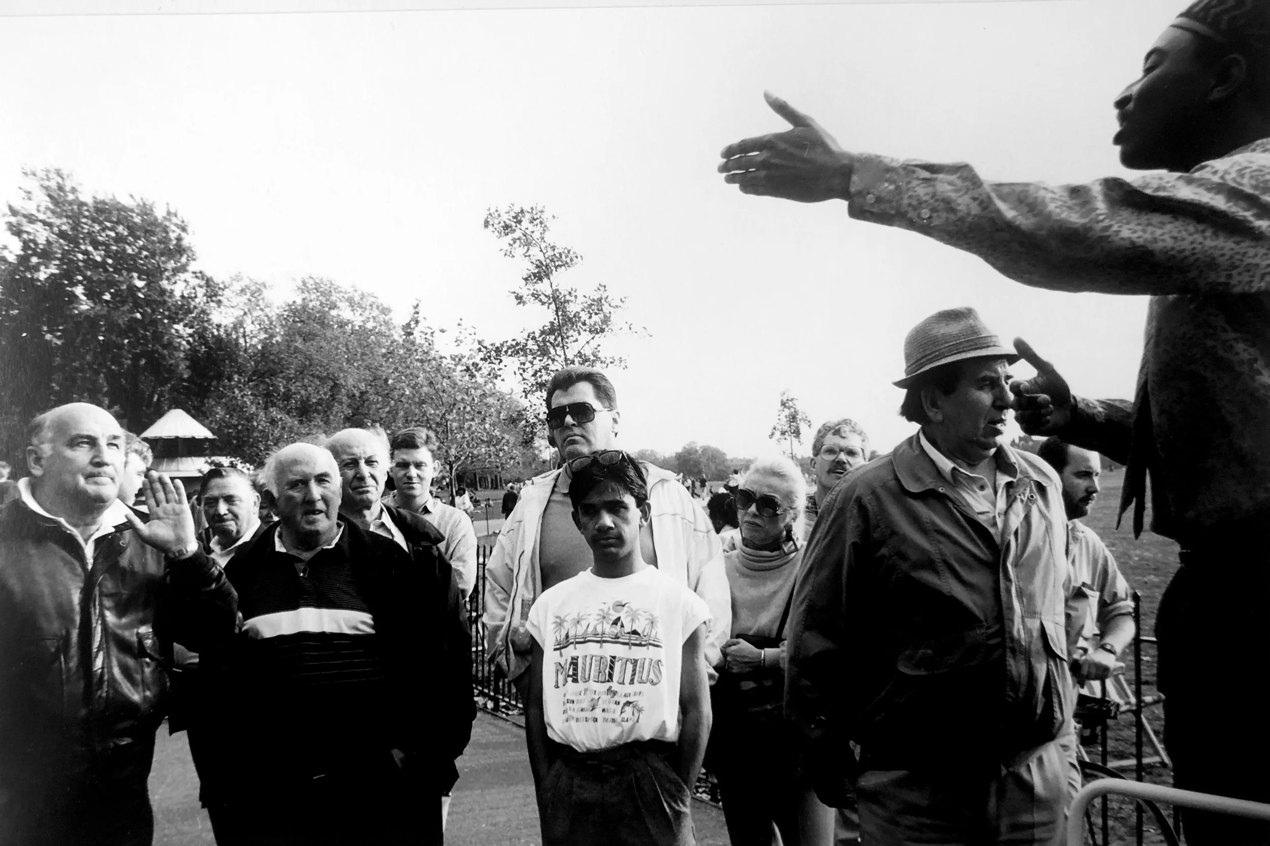 A group of people gathered outdoors, with a woman gesturing and speaking to them. The group includes men of different ages, some with sunglasses, and one wearing a hat. Trees and a cloudy sky are visible in the background.