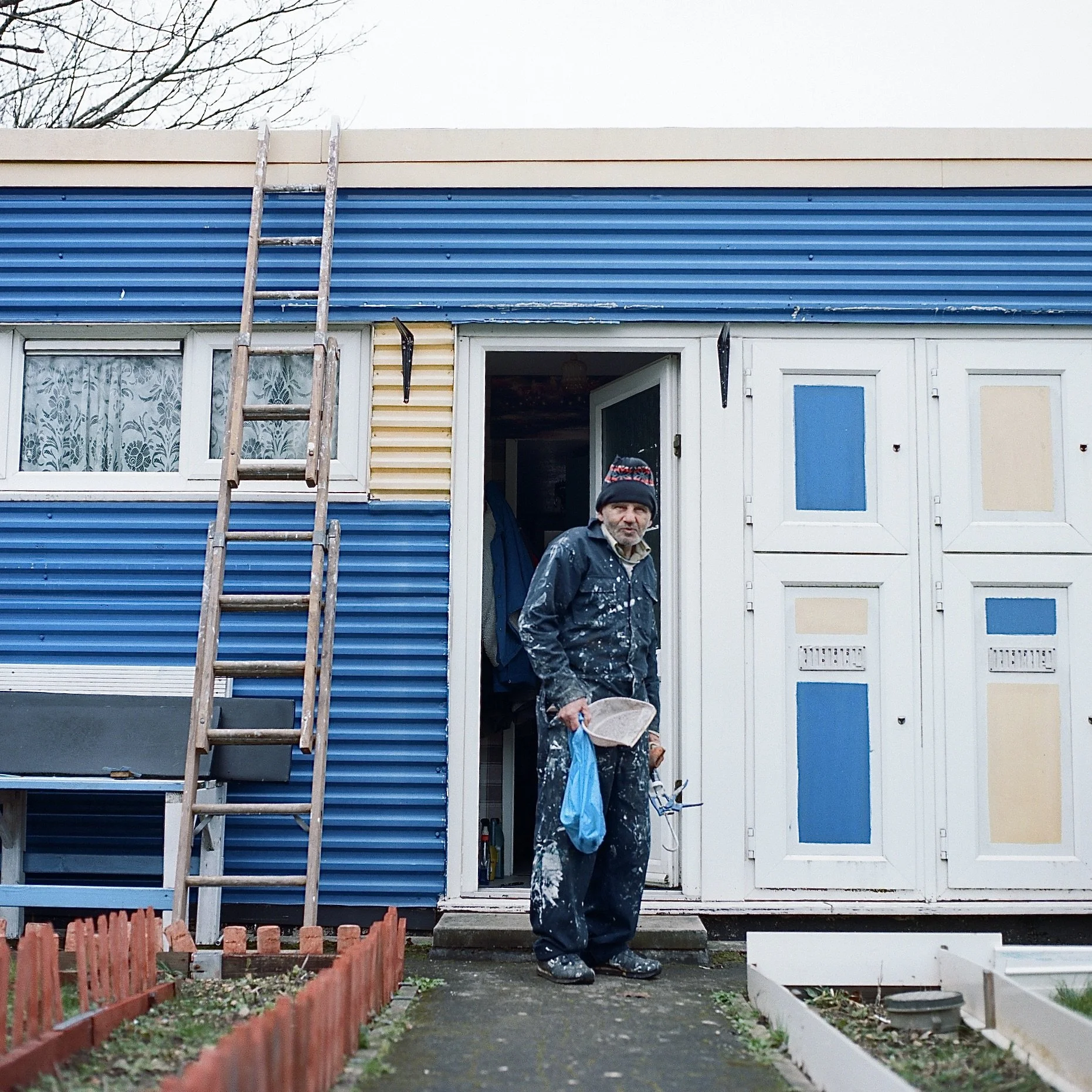 A man wearing a black jacket with white paint splatters, black pants with white paint splatters, black boots, and a black cap stands in front of a house with a blue and beige exterior. The house has a ladder leaning against the roof, and a partially 