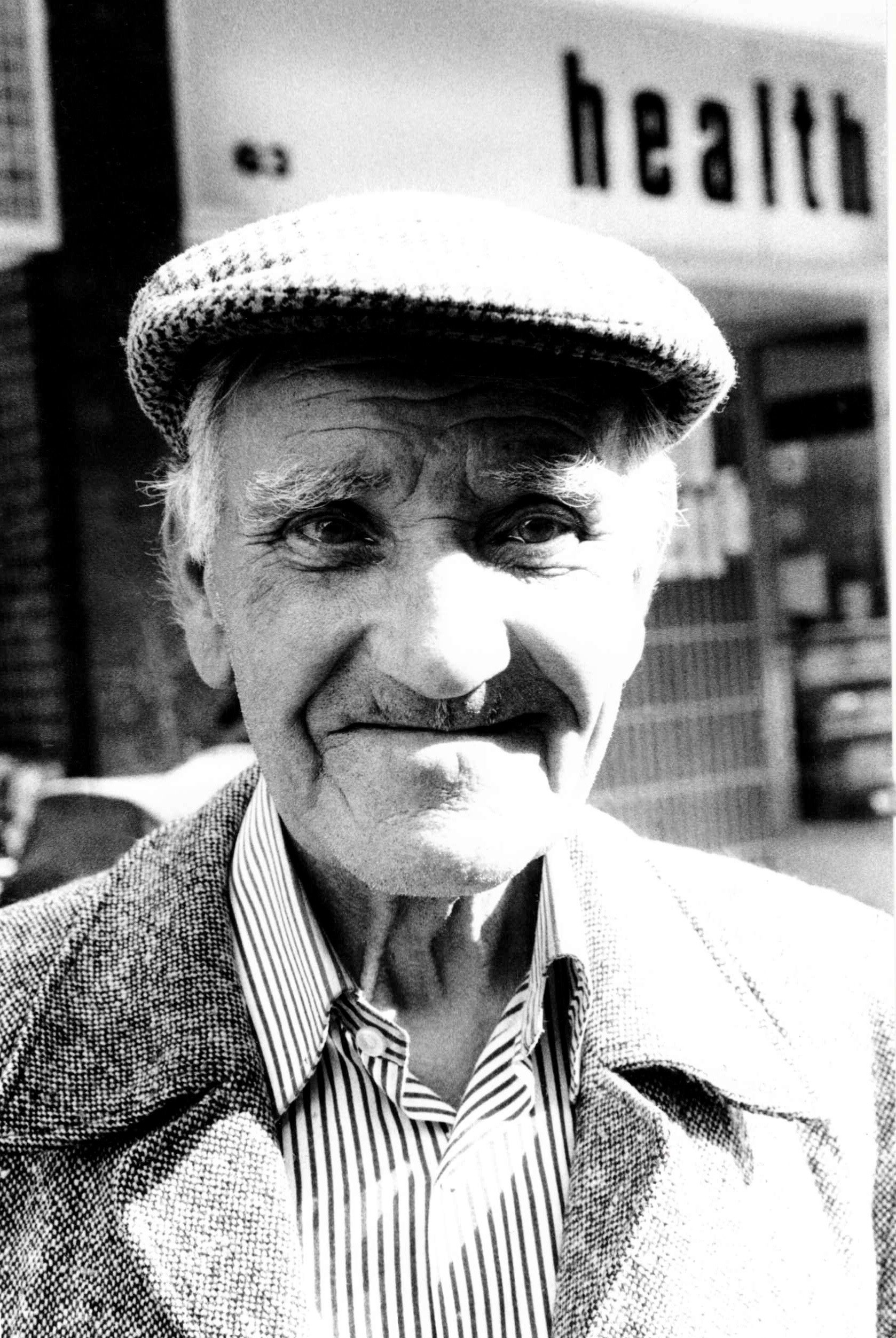 Close-up black and white photo of an elderly man wearing a checkered blazer, striped shirt, and cap, smiling outdoors, with the word 'health' partially visible in the background.