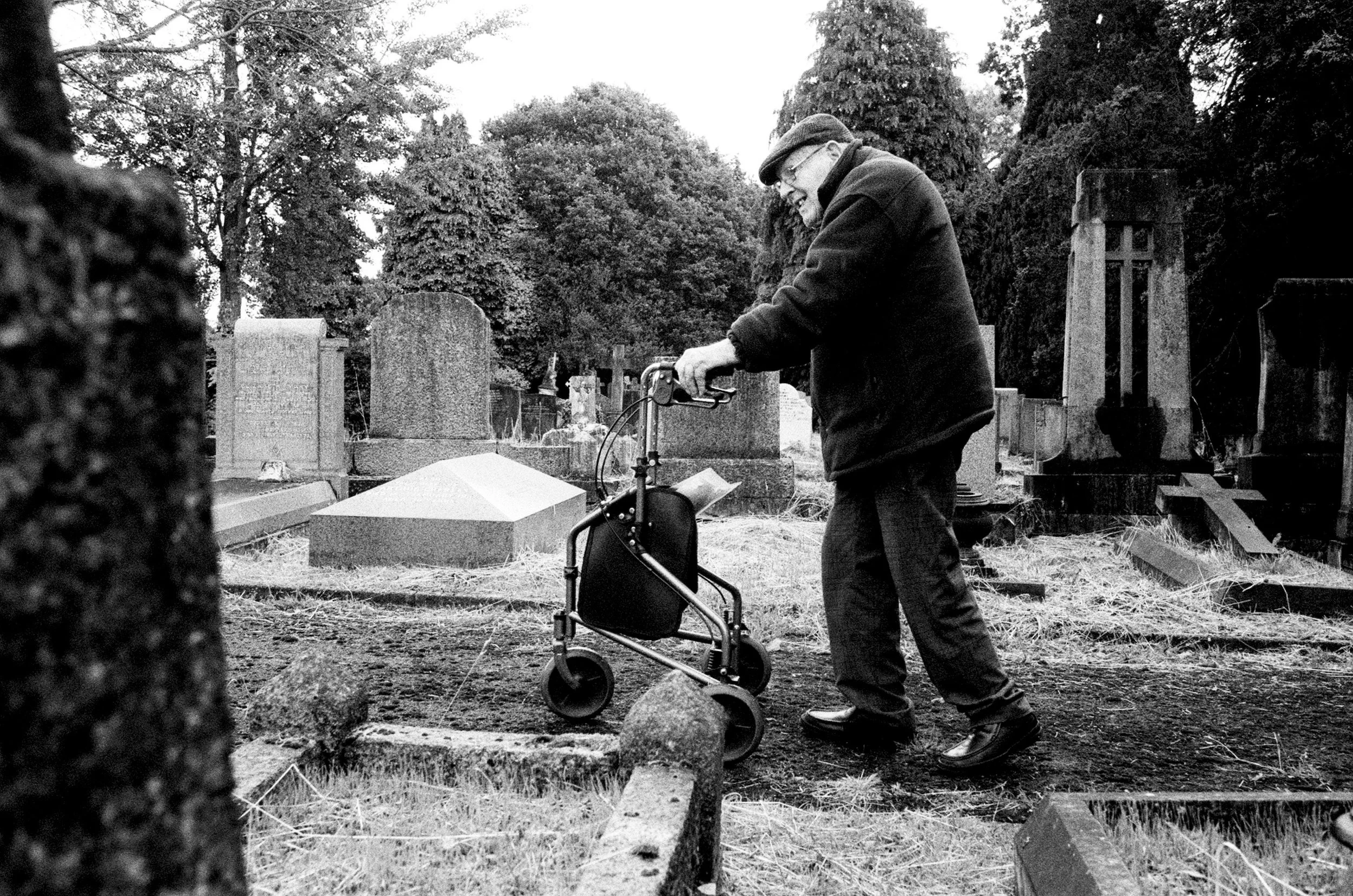 An elderly man smiling while walking with a walker in a cemetery, surrounded by tombstones and trees.