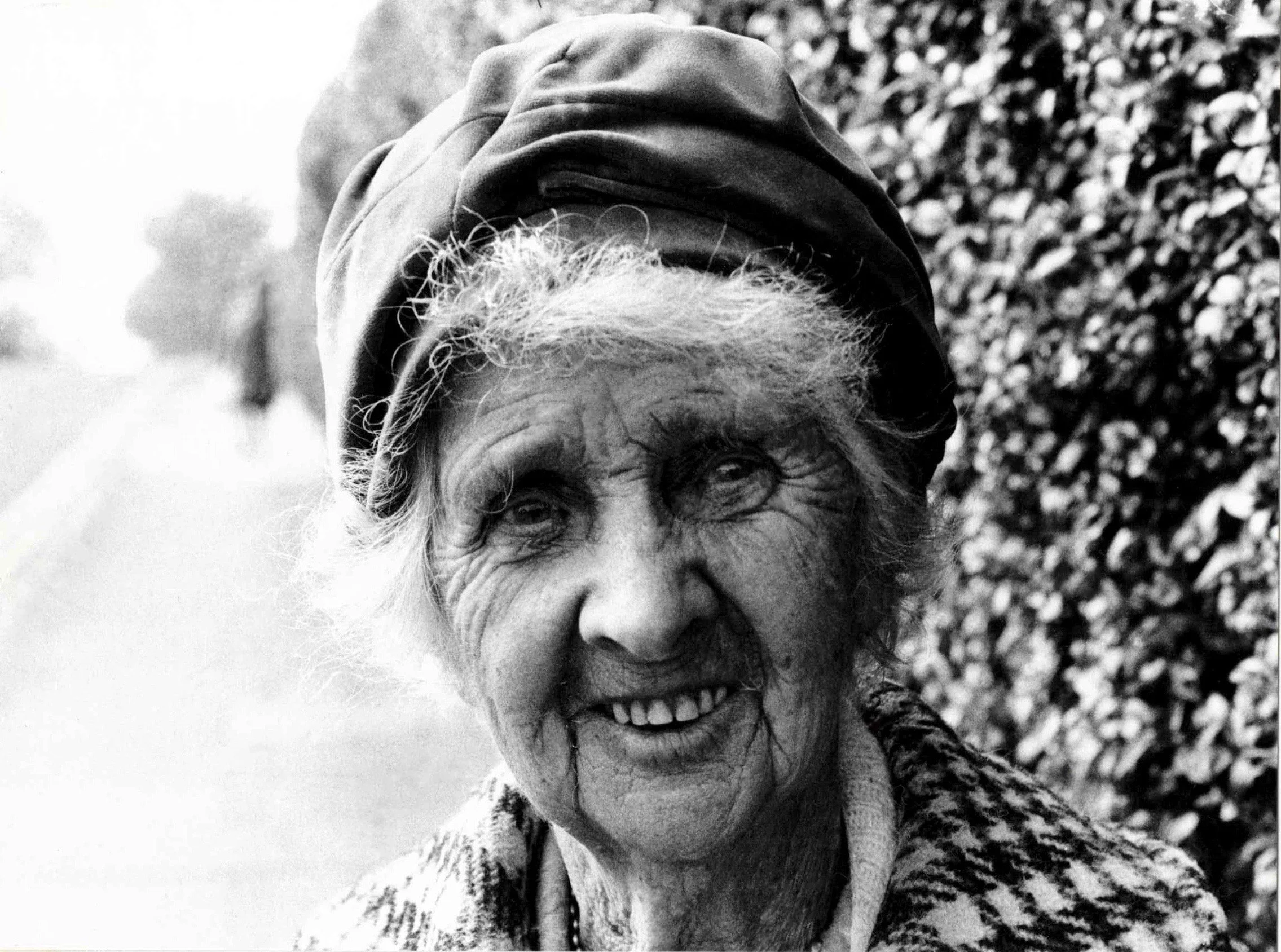 A smiling elderly woman with curly hair wearing a headscarf outside, with a blurred shrub or bush in the background.