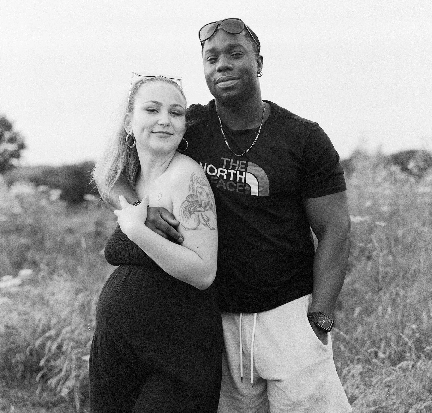 A black and white photo of a young couple outdoors. The woman has light skin, long hair, hoop earrings, and a tattoo on her shoulder. The man has dark skin, short hair, sunglasses on his head, and is wearing a The North Face T-shirt. They are hugging