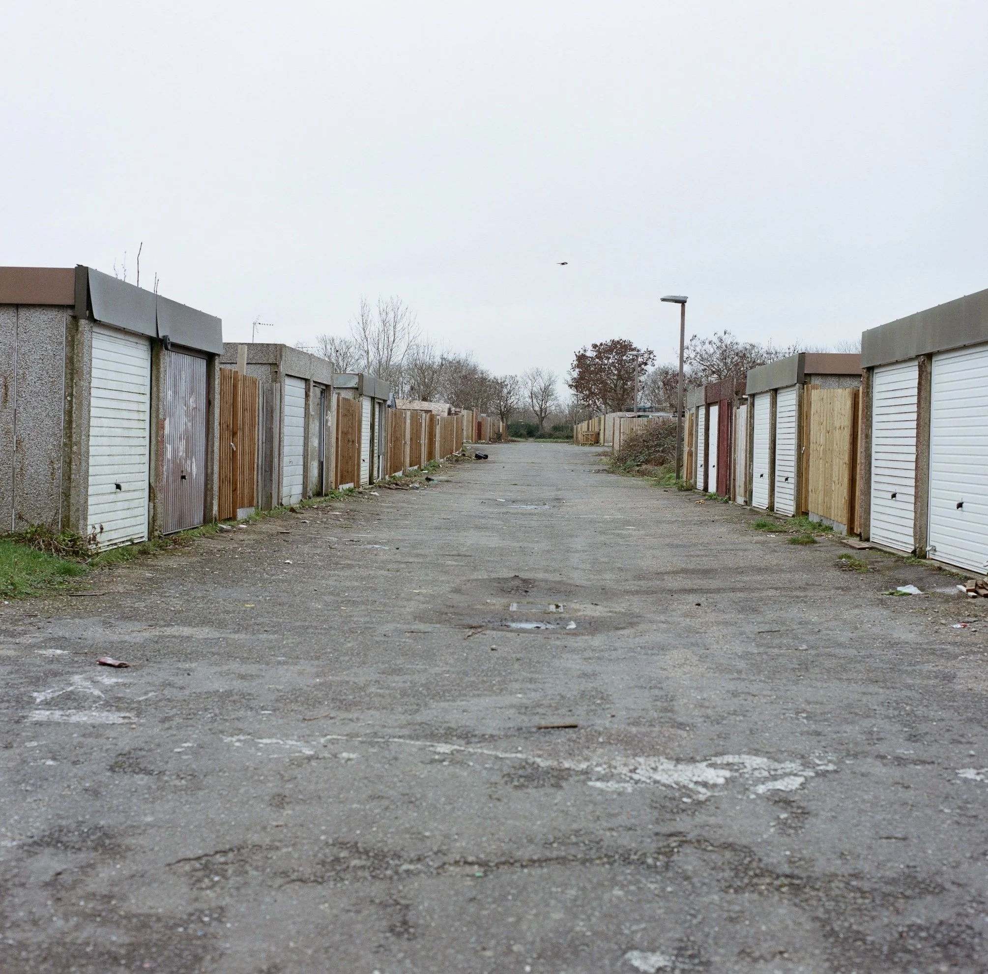 An empty alleyway lined with garages on both sides, with trees and street lamps visible in the background, under an overcast sky.