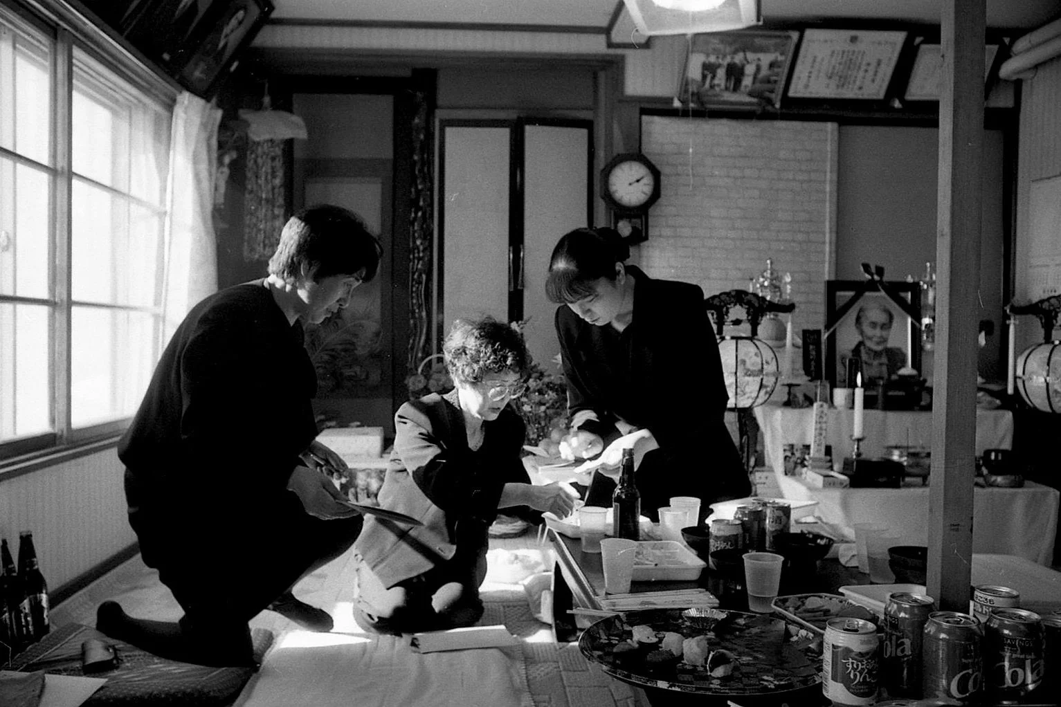 Three people indoors, one elderly woman seated, and two younger women standing and kneeling, examining papers or books on a table, with food and drink cans nearby, in a room with a clock and various decorations.