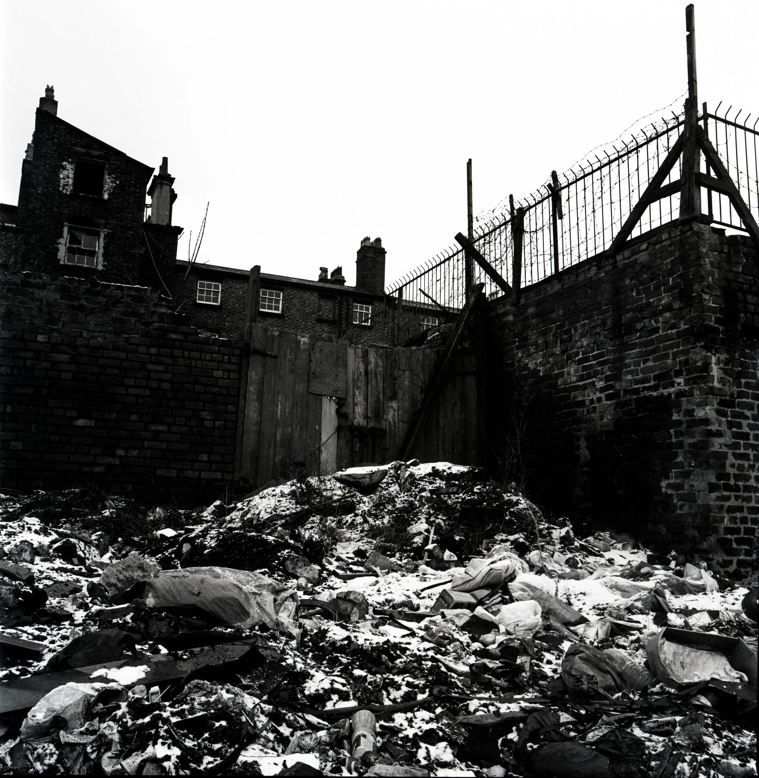A black and white photo of a dilapidated building with broken windows and a barbed wire fence on top of a brick wall, with debris and garbage scattered on the ground in front.