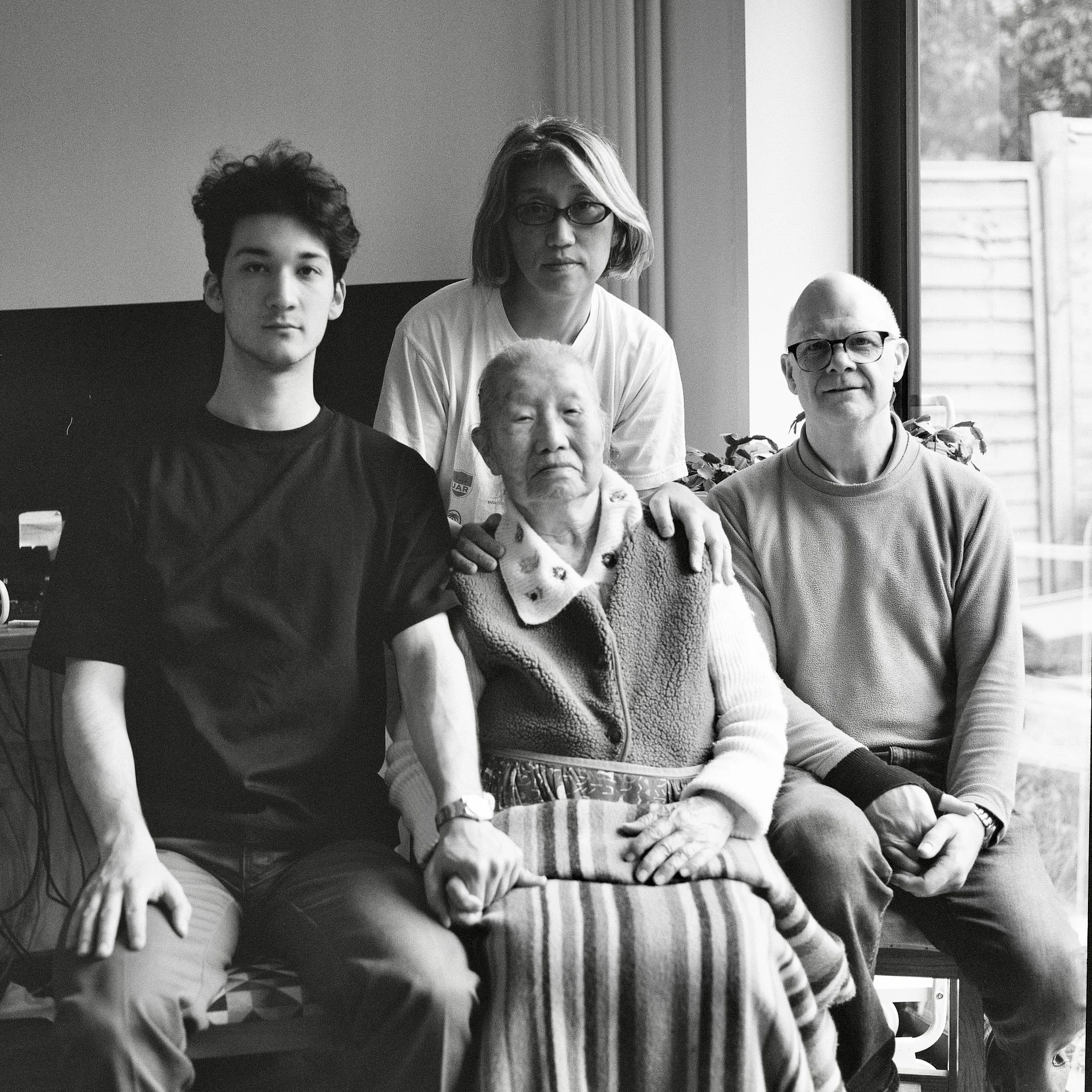 A black and white photograph of a family of five sitting together indoors near a large window, with a view of an outdoor fence. The family includes a young man, an older woman, an elderly woman, an elderly man, and a middle-aged man. They are dressed