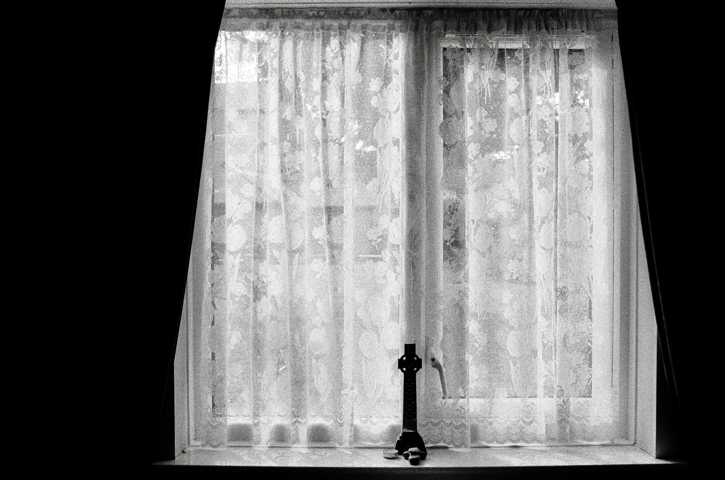 A black and white photo of a window with curtains. There are objects on the windowsill and lace curtains hanging in front of the window.