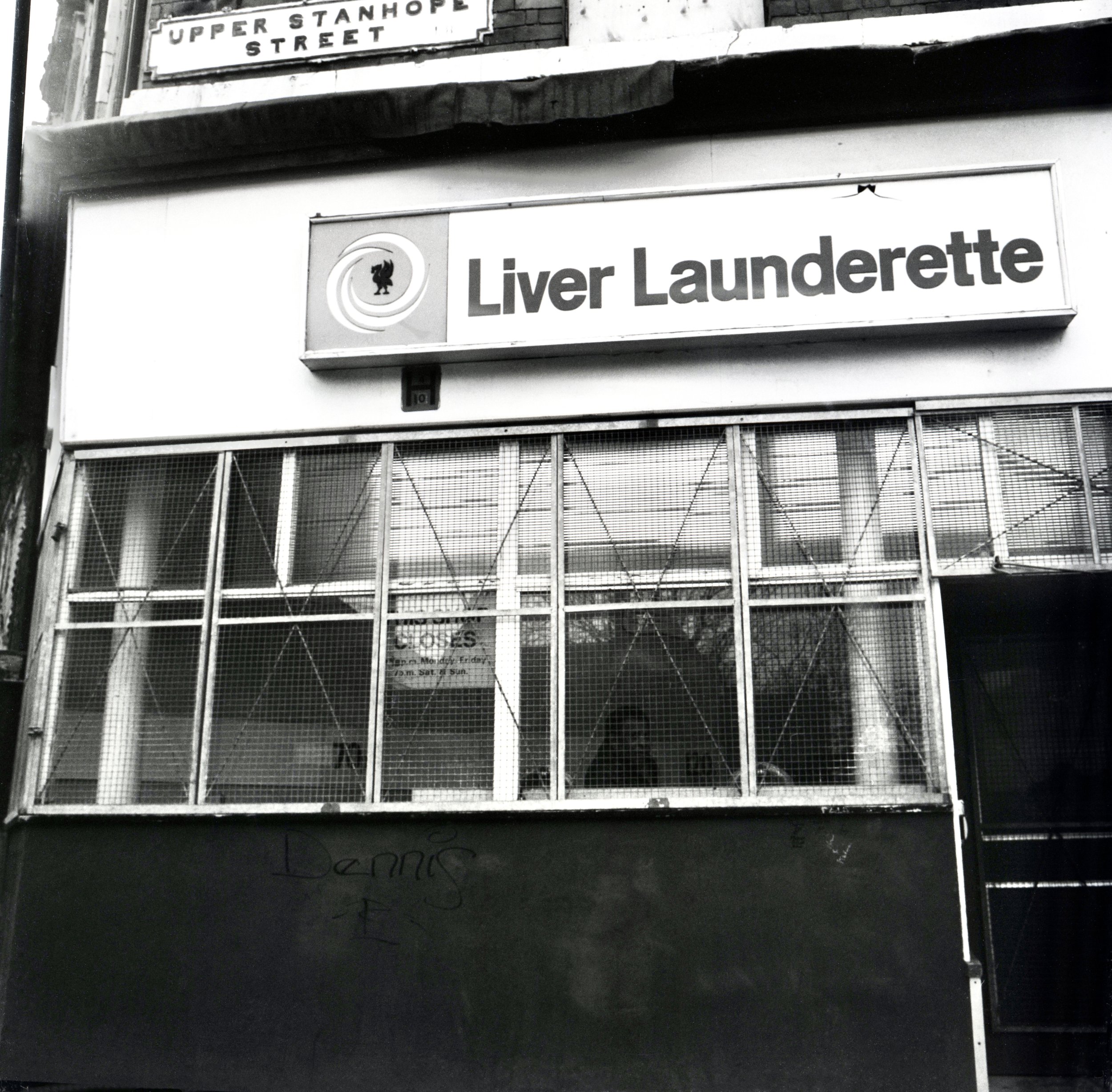 Black and white photo of a street corner with a sign for Upper Stanhope Street and a sign for Liver Launderette, which features a circular logo with a silhouette of a person or figure and text on a storefront window, with a metal gate or mesh below.