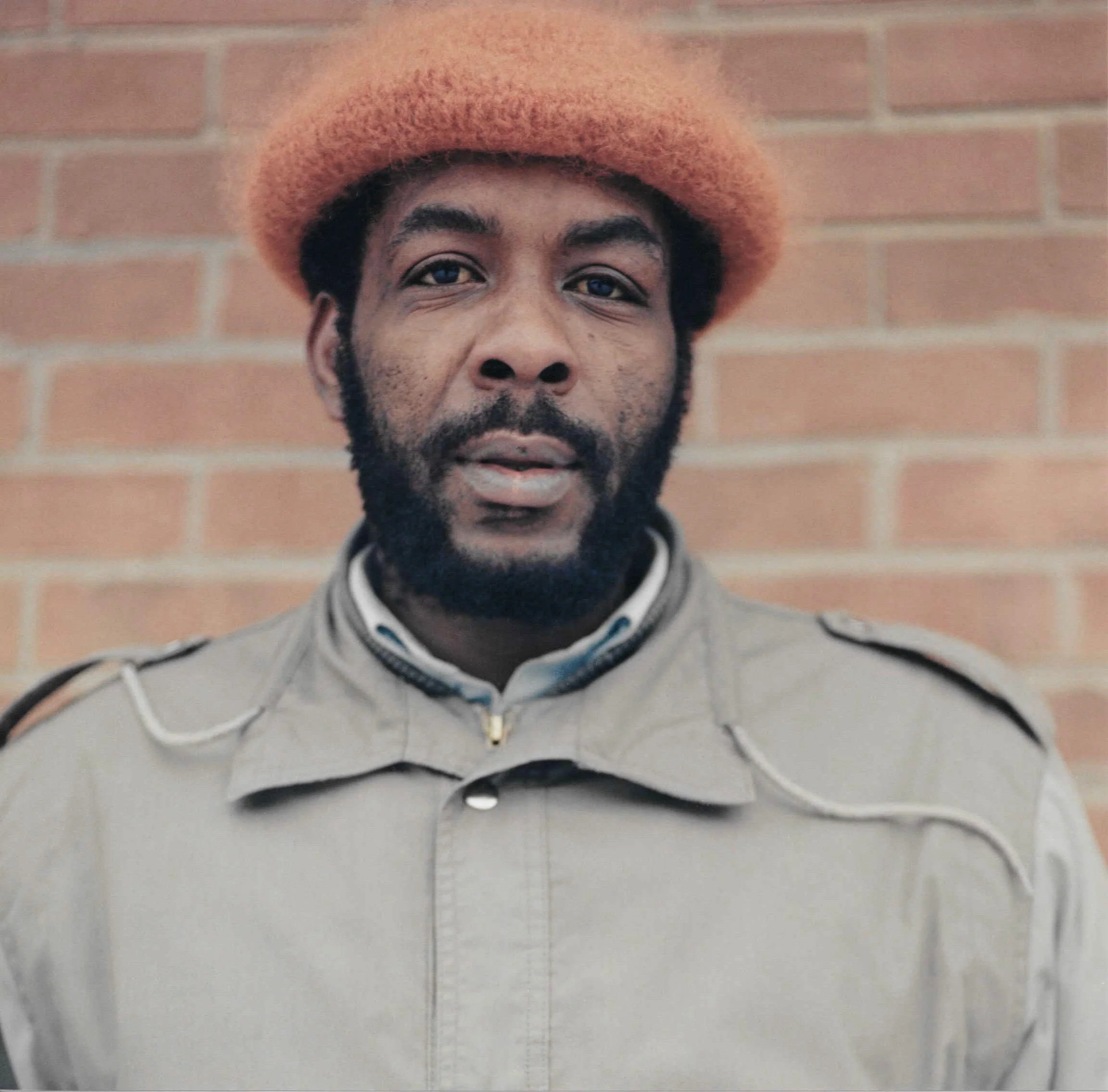 A person with a beard and mustache wearing a light-colored hoodie, earbuds, and an orange hat, standing in front of a brick wall.